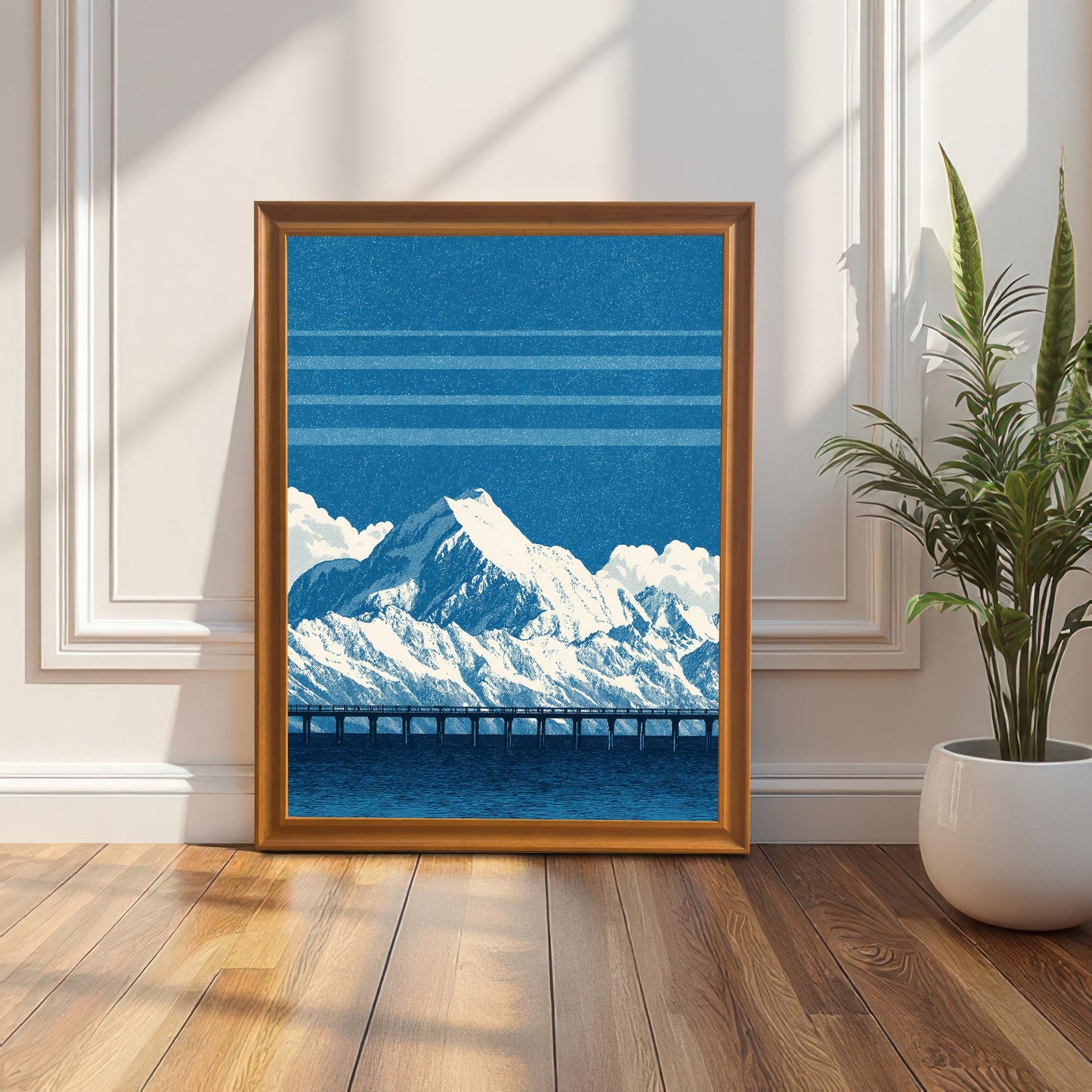 A framed art print of the blue mountain and bridge design in an Orange (golden/bronze) frame, leaning against a white wainscoting wall next to a potted plant, with sunlight streaming across the wooden floor.