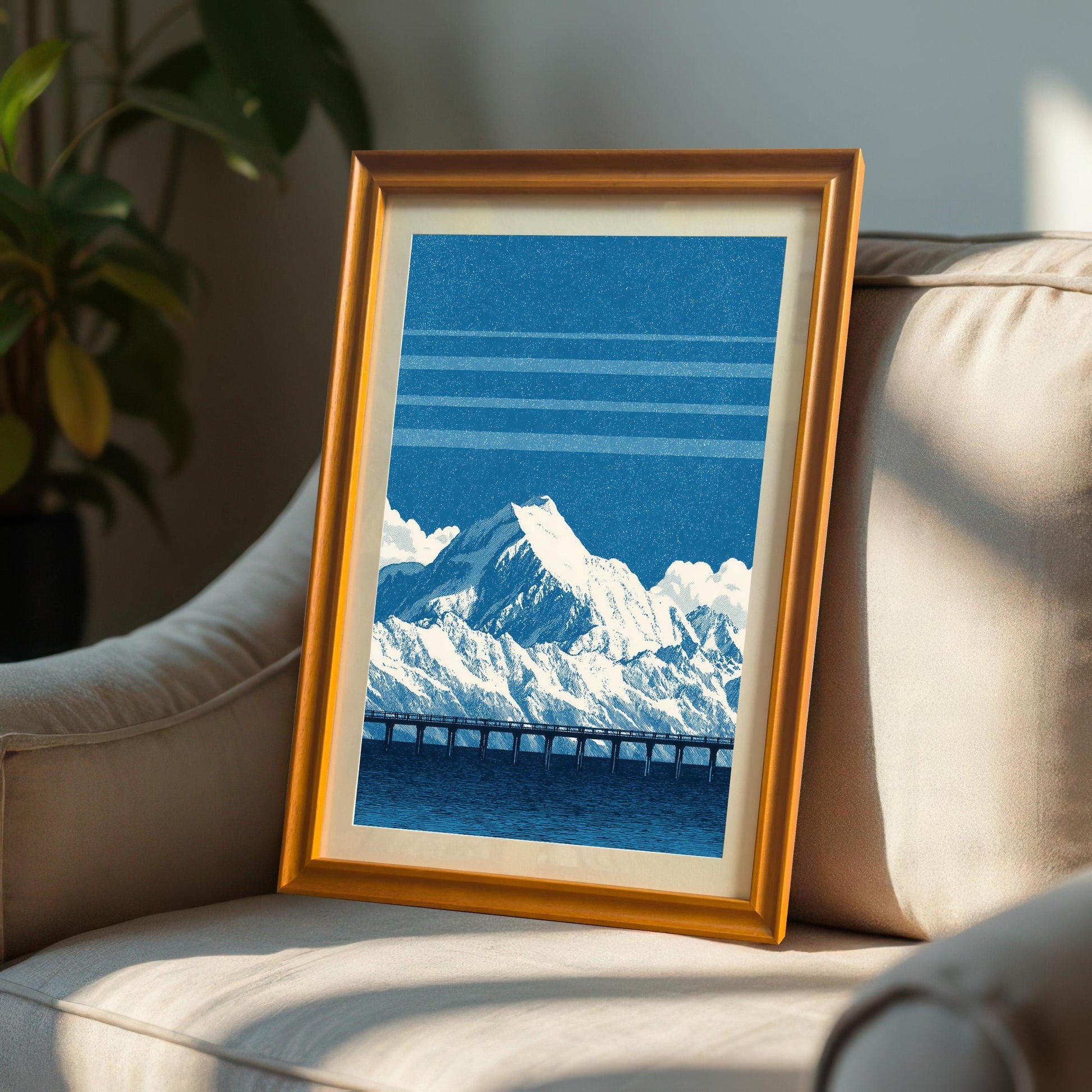 A framed art print of the blue mountain and bridge design in an Orange (golden/bronze) frame, leaning on a light-colored armchair or sofa, with sunlight hitting the frame.