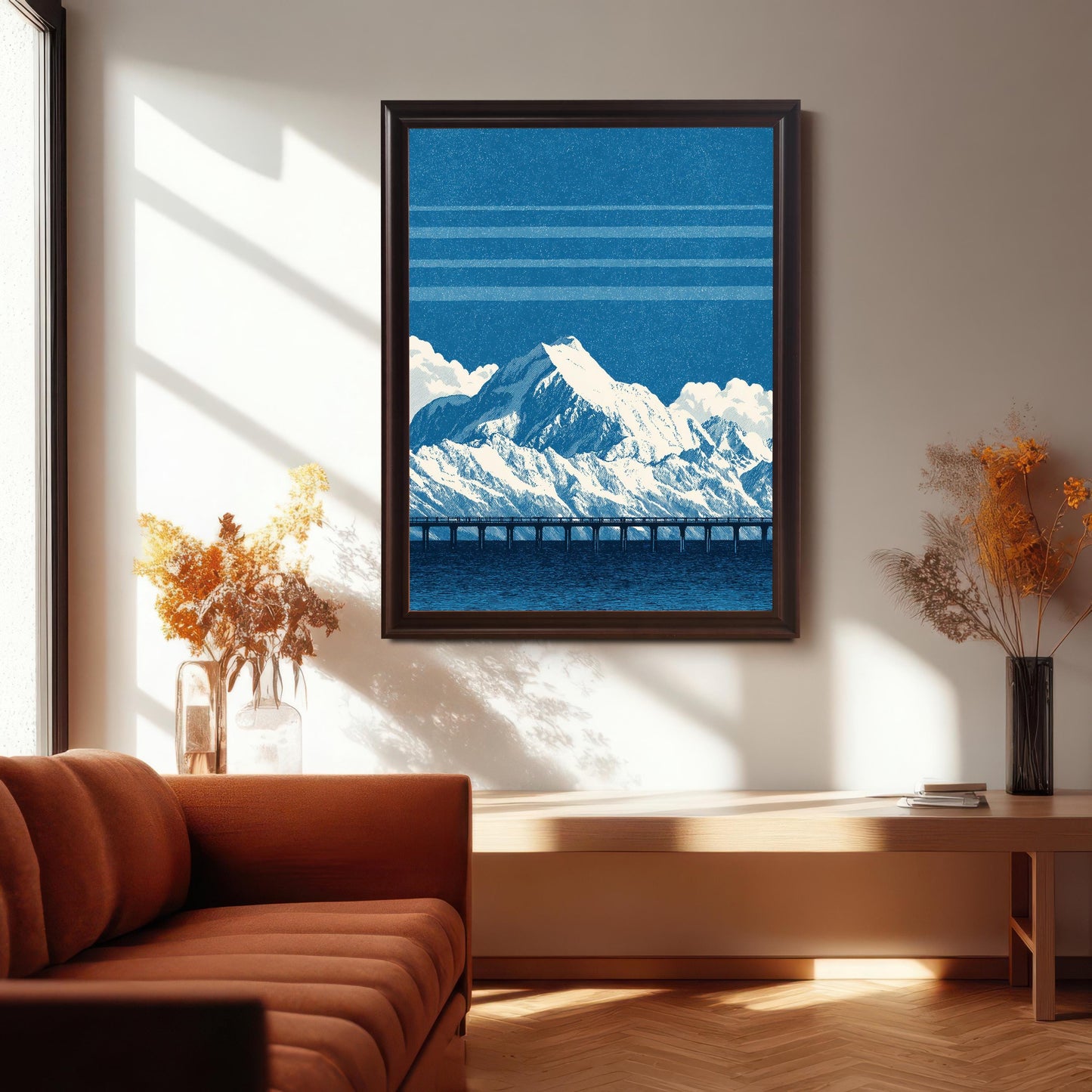 The blue mountain and bridge art print in a dark brown/coffee frame, hanging on a white wall above a wooden console table. The room features a terracotta orange sofa, vases with dried flowers, and is brightly lit by sunlight streaming through a large window.