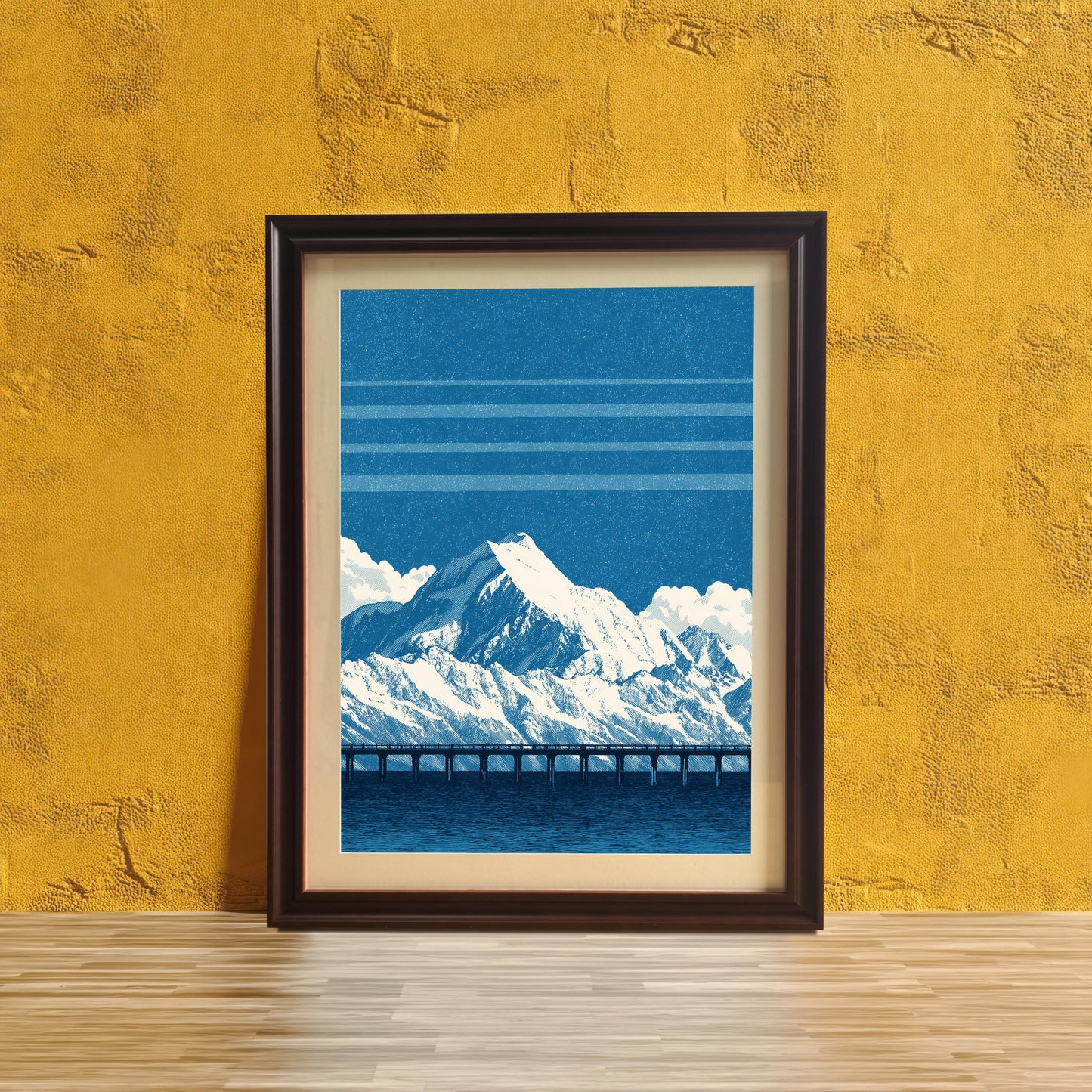 The blue mountain and bridge art print in a dark brown frame with a mount/mat, leaning on a light wooden surface against a heavily textured, vibrant yellow wall.