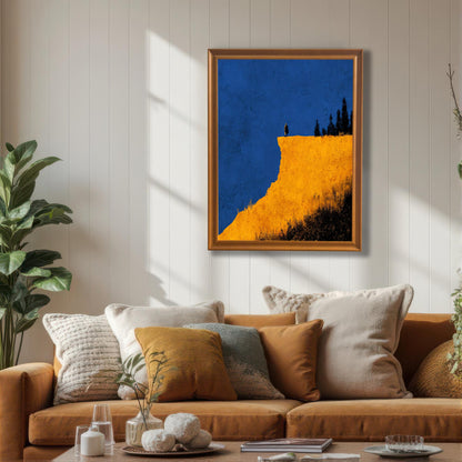 Abstract art print of a figure on a yellow cliff against a blue sky, in an Orange/light brown wood frame, hanging above a tan sofa with warm-toned pillows in a living room. Shows the print used in a mid-century modern or bohemian setting.