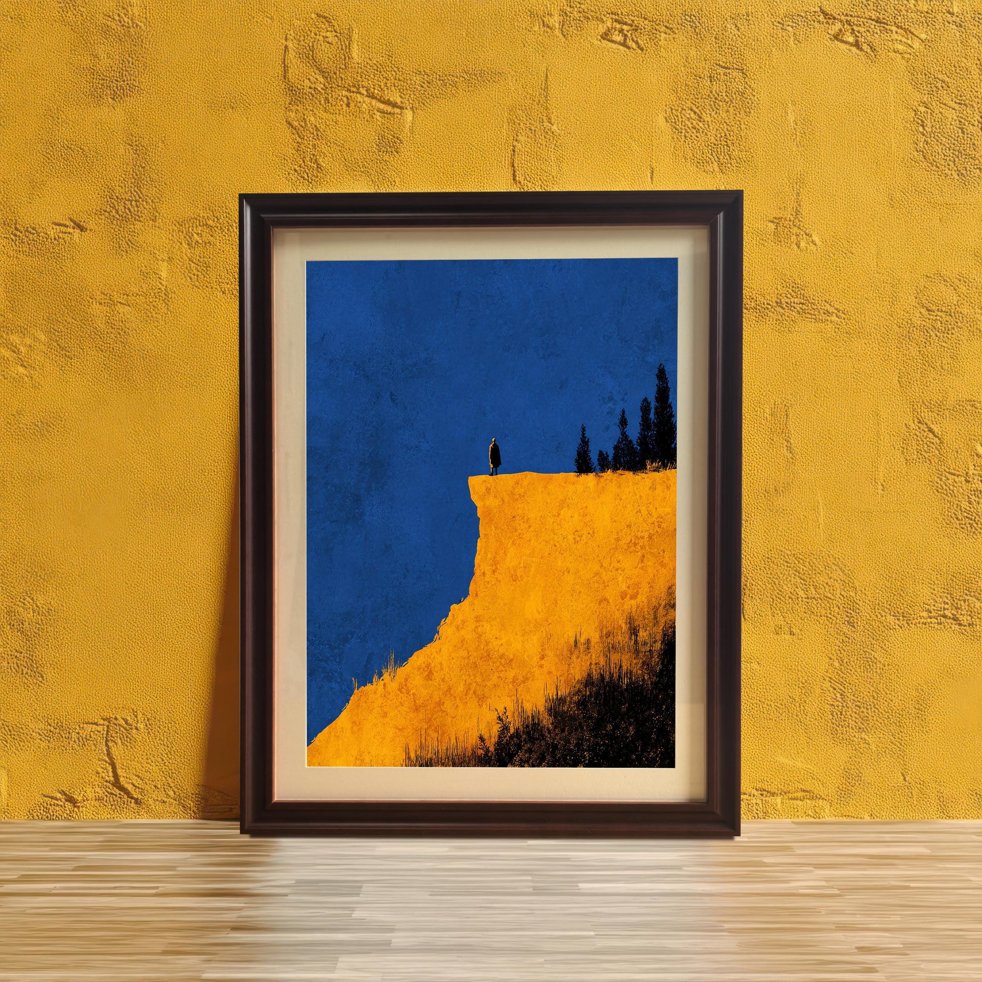 Abstract cliff print in a Coffee (dark brown) frame with a mount, leaning against a vibrant yellow textured wall on a wooden surface. Features bright yellow cliff and deep blue sky.