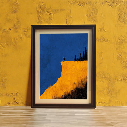 Abstract cliff print in a Coffee (dark brown) frame with a mount, leaning against a vibrant yellow textured wall on a wooden surface. Features bright yellow cliff and deep blue sky.