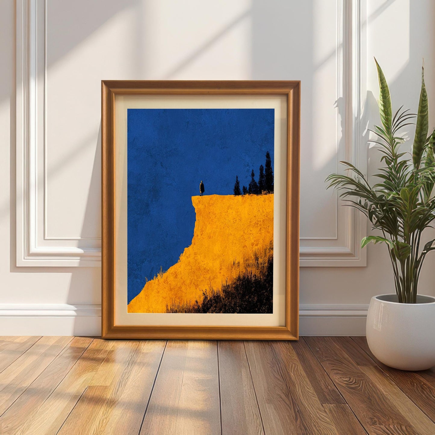 Abstract art print of a figure on a dramatic yellow cliff against a blue sky, in an Orange/light brown wood frame with a mount. The print is leaning on a wooden floor against a white paneled wall, next to a tall snake plant in a white pot.
