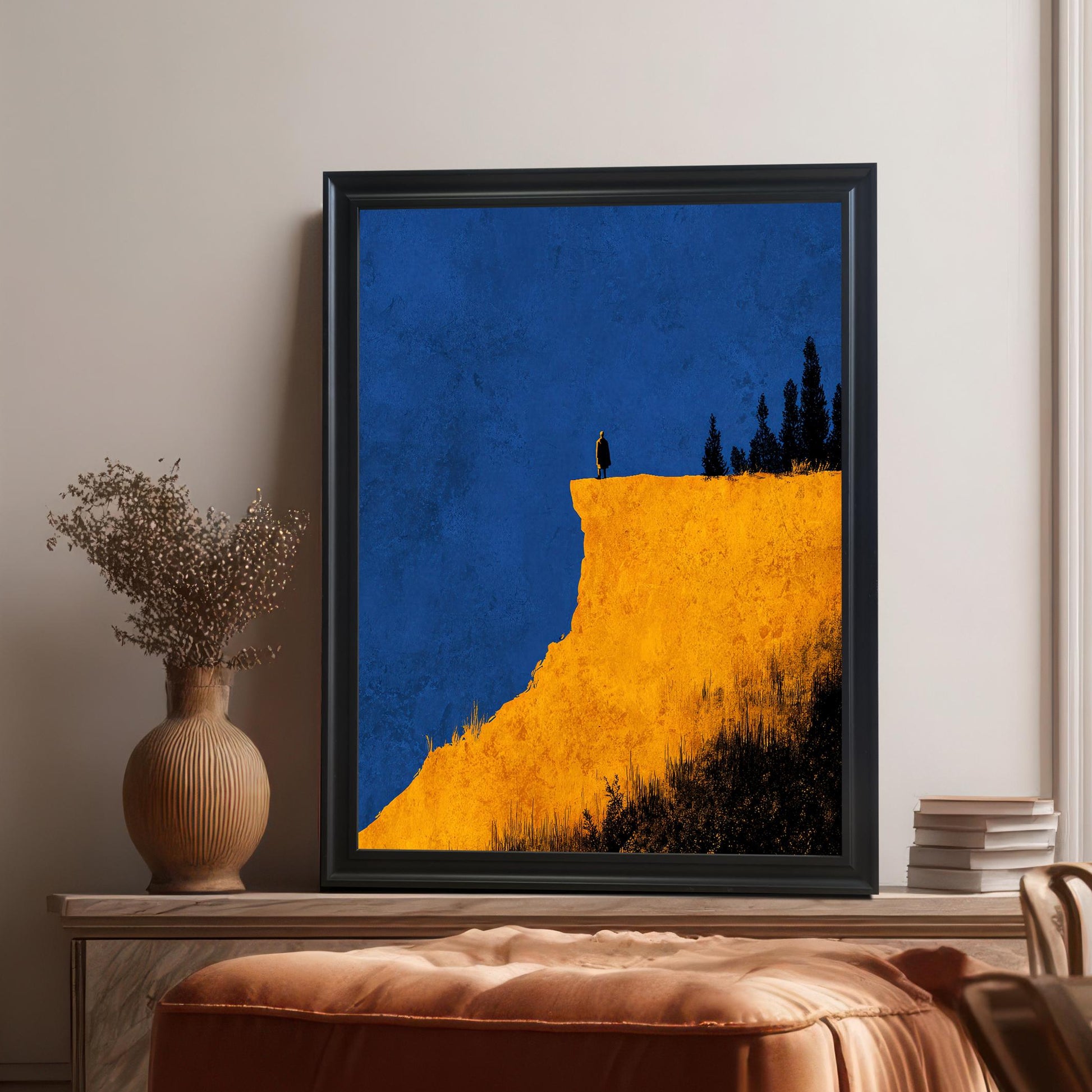 Abstract yellow cliff print in a Black frame (no mount), leaning on a soft brown leather ottoman. A vase with dried flowers is on the left. Shows the print in a modern interior.