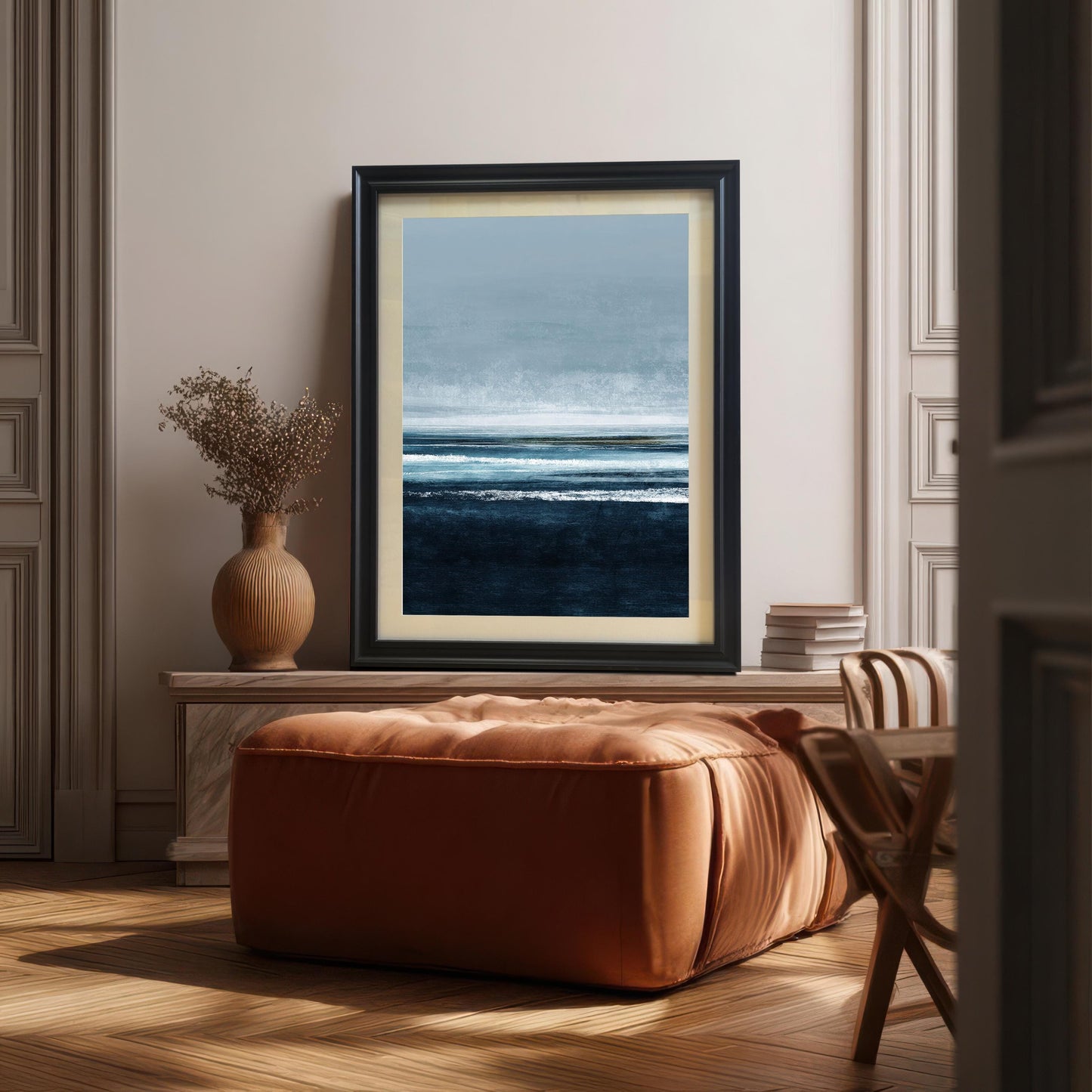 Abstract seascape art print in a Black frame with a mount/mat, placed on a white console table. A large brown leather ottoman is in the foreground, and a brown vase with dried foliage is on the left. The scene is stylish and contemporary.