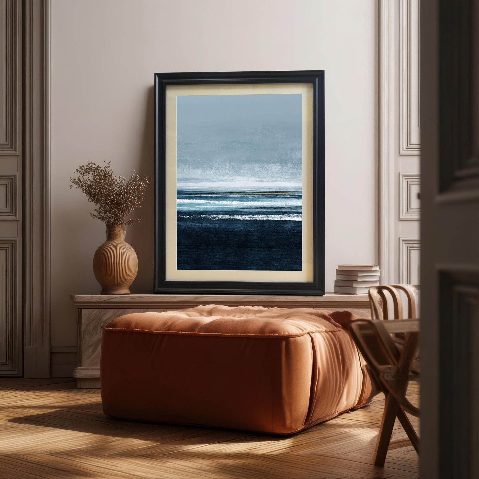 Abstract seascape art print in a Black frame with a mount/mat, placed on a white console table. A large brown leather ottoman is in the foreground, and a brown vase with dried foliage is on the left. The scene is stylish and contemporary.