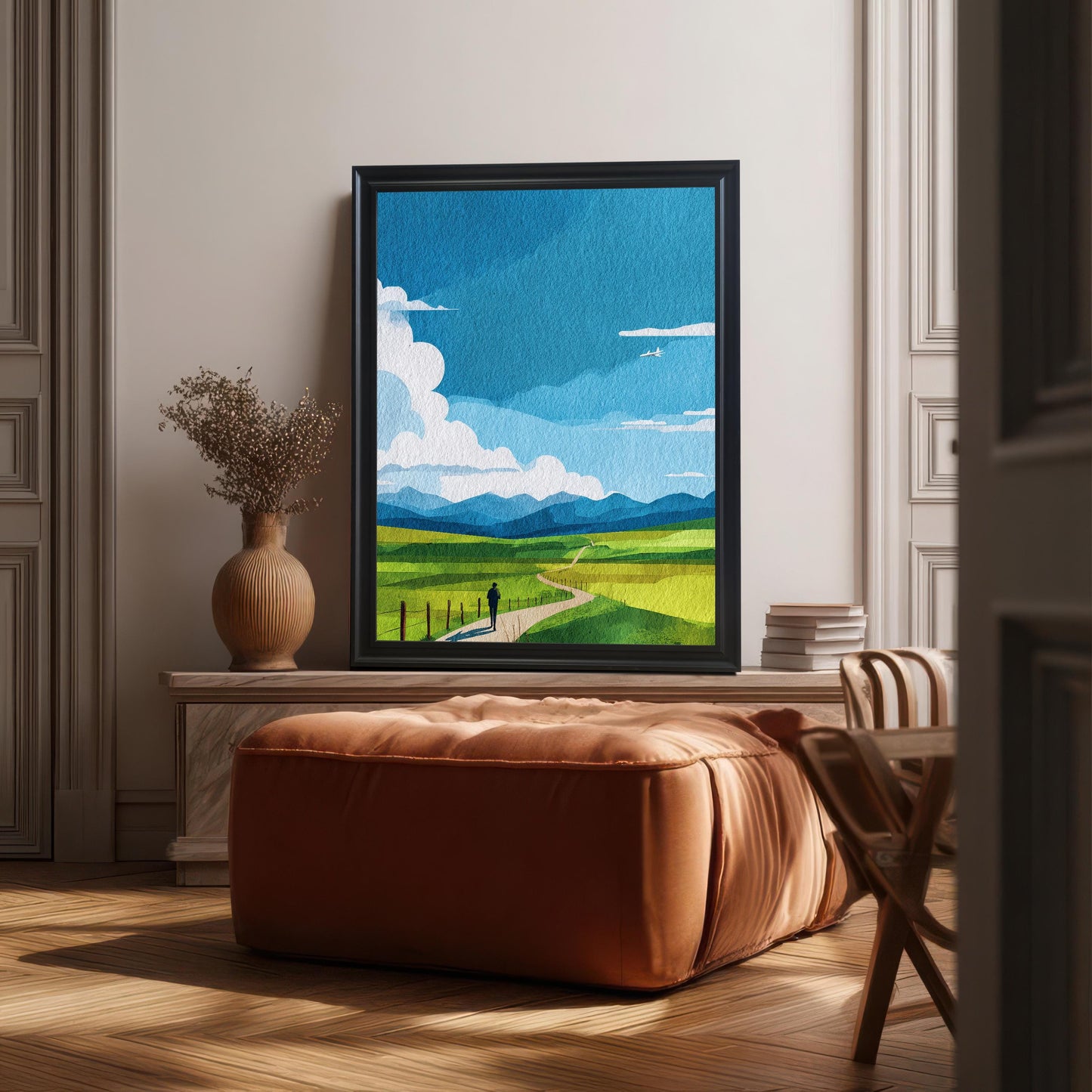 Art print of the blue sky and green field landscape in a Black frame (no mount), placed on a white console table. A large brown leather ottoman is in the foreground, and a terracotta vase with dried flowers is on the left. The scene is illuminated by strong sunlight and shadows.