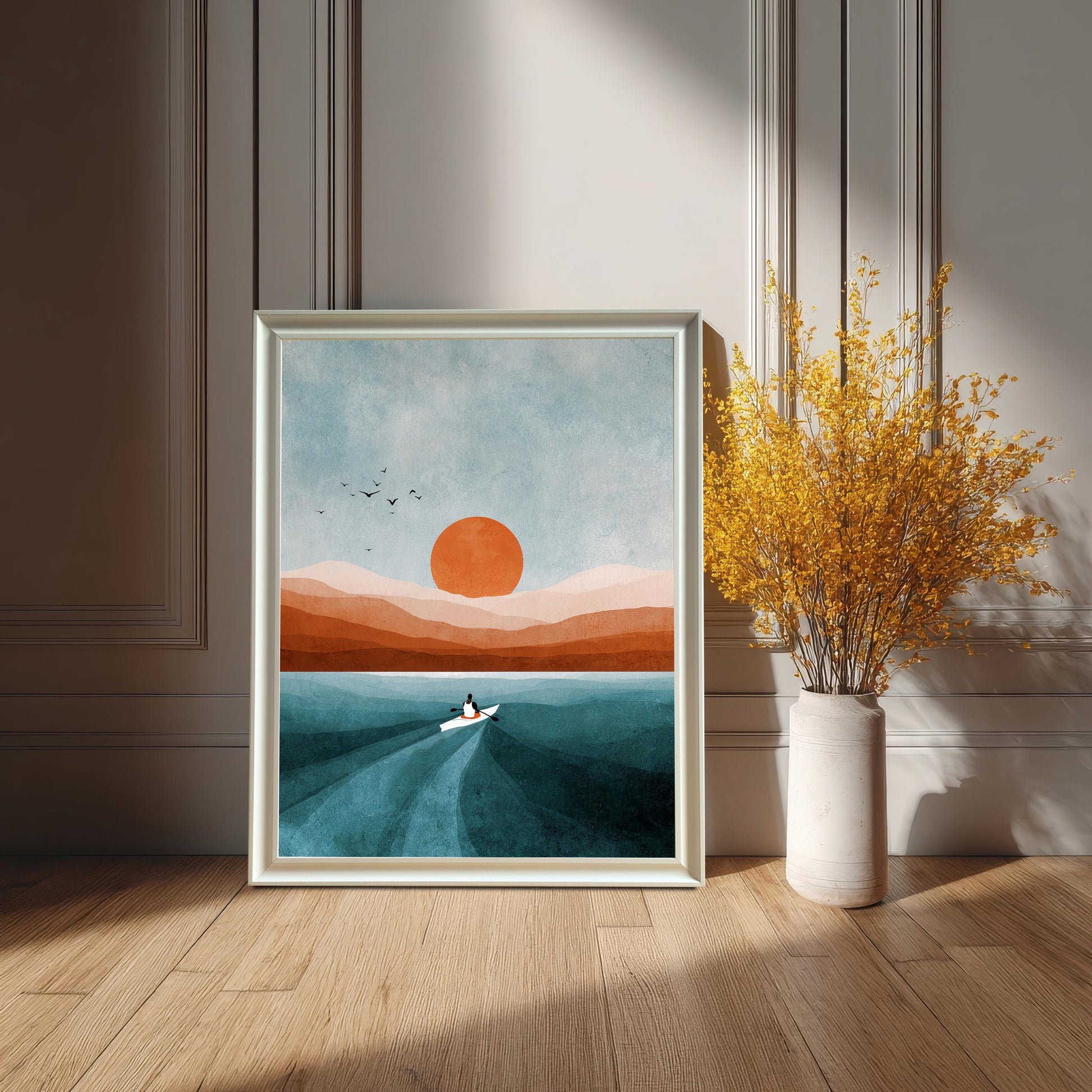 White-framed art print leaning on a wooden floor against a white paneled wainscoting wall, featuring a modern abstract landscape with a boat, orange sun, and teal water. A tall white vase with bright yellow dried flowers is beside it.