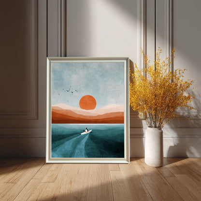 White-framed art print leaning on a wooden floor against a white paneled wainscoting wall, featuring a modern abstract landscape with a boat, orange sun, and teal water. A tall white vase with bright yellow dried flowers is beside it.