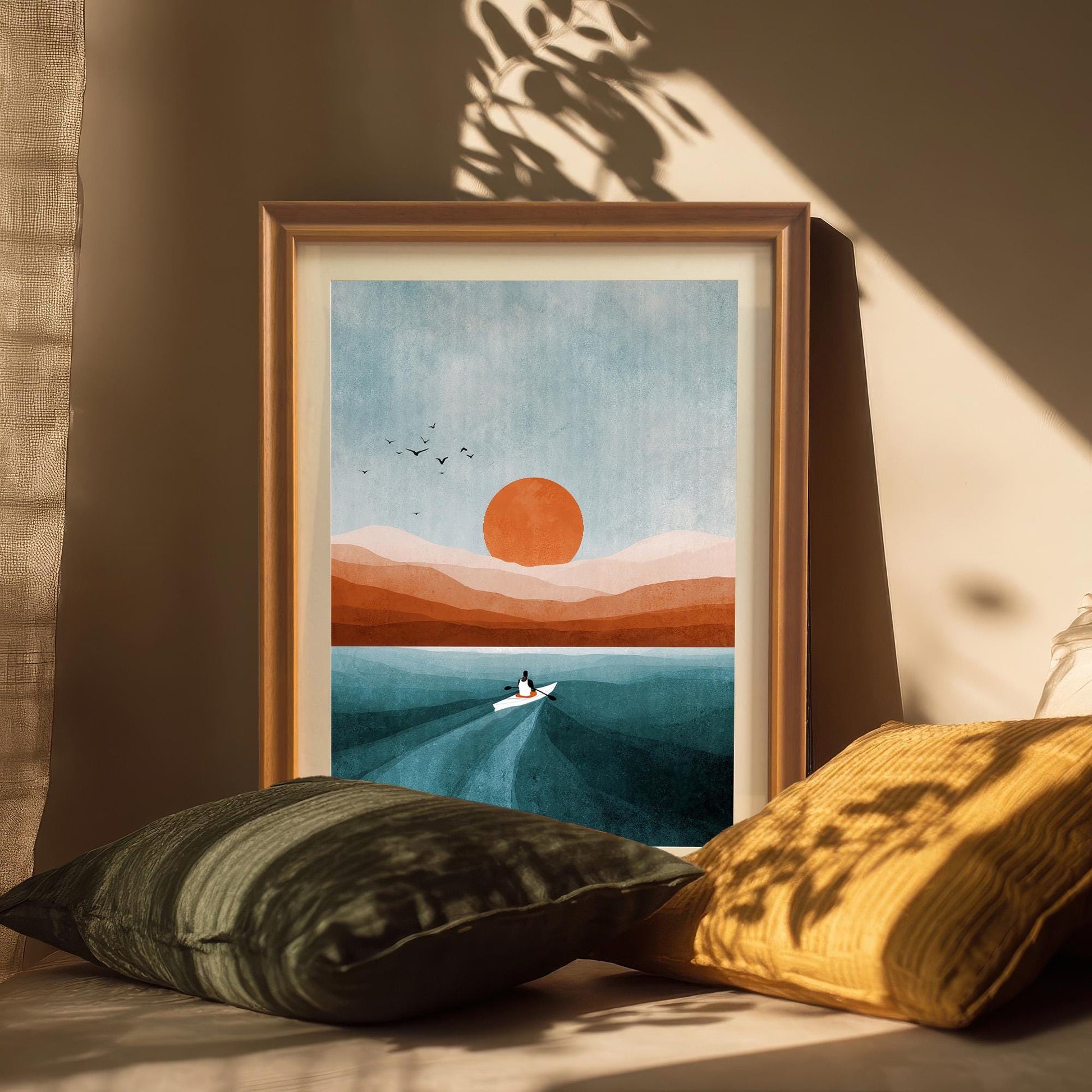 Abstract sunset/sunrise print of a lone person in a boat on teal blue water with orange hills and sun, displayed in a wood frame leaning against a tan wall, with sunlight and shadows, and two velvet throw pillows (green and gold). Ideal for bohemian or minimalist decor.