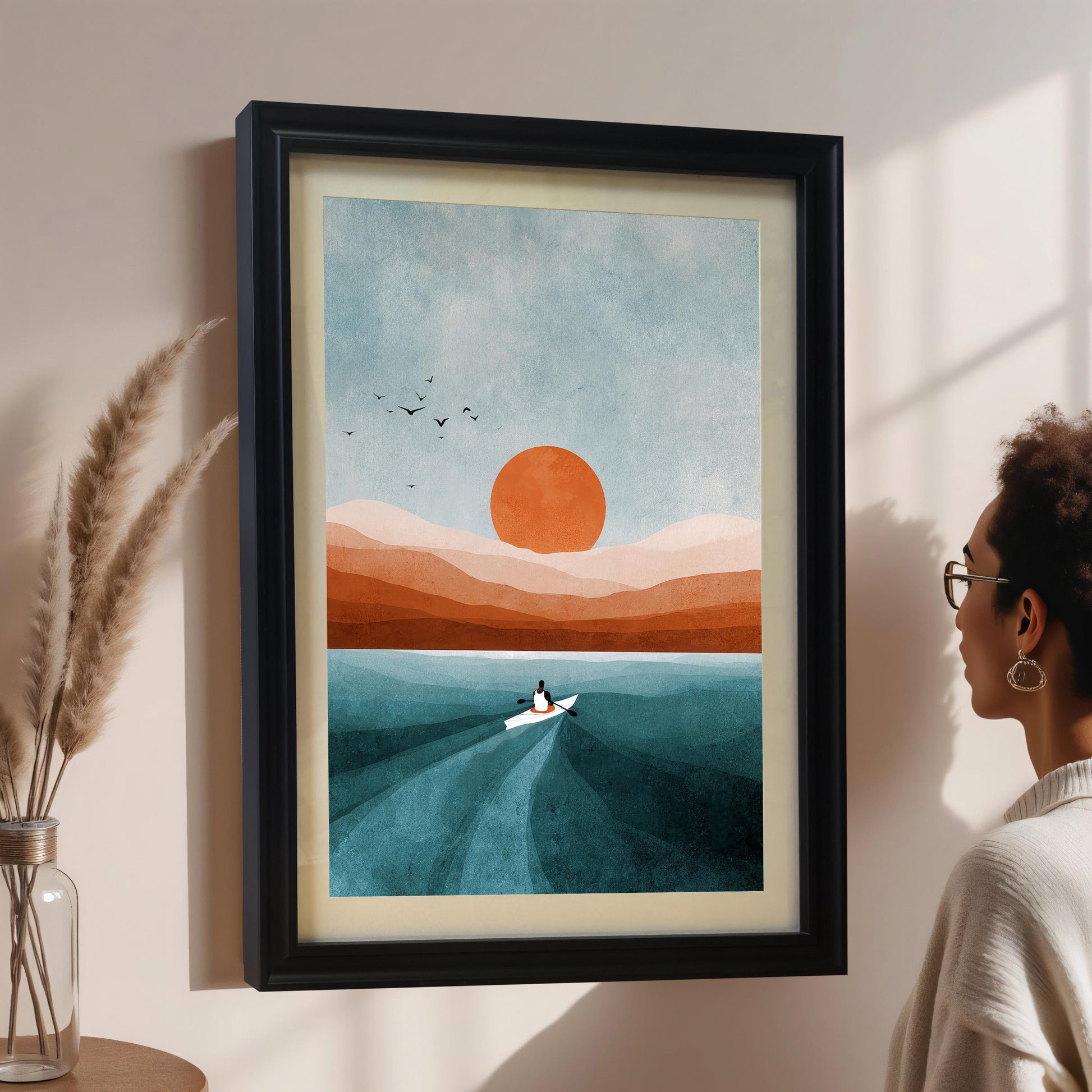A woman in glasses and a light sweater looks at a framed abstract art print on a pale pink wall. The print features a boat, orange sun, and blue/orange landscape, displayed in a black wood frame with a cream-colored mount/mat. Pampas grass in a clear vase is on the left.
