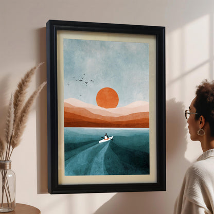A woman in glasses and a light sweater looks at a framed abstract art print on a pale pink wall. The print features a boat, orange sun, and blue/orange landscape, displayed in a black wood frame with a cream-colored mount/mat. Pampas grass in a clear vase is on the left.