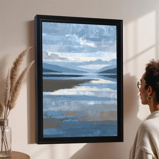 A woman with glasses and curly hair looking at the abstract blue and brown landscape print in a black frame, which is hanging on a light wall next to a vase of pampas grass.