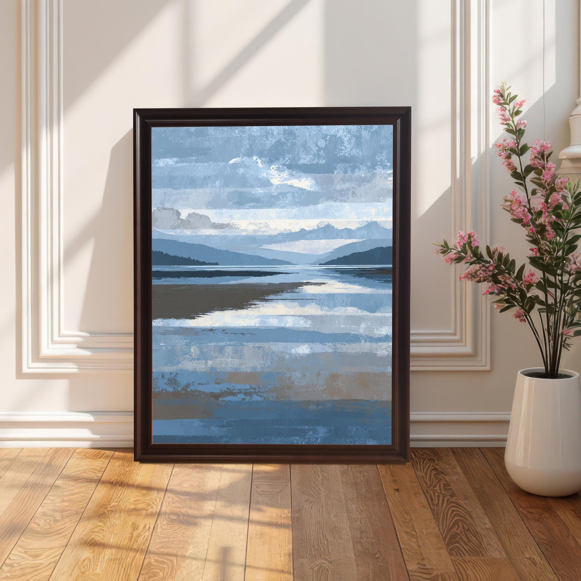 Abstract blue and brown landscape art print framed in dark Coffee wood with a light mount, leaning against a white wainscoted wall on a wooden floor, next to a tall white vase with pink flowers.