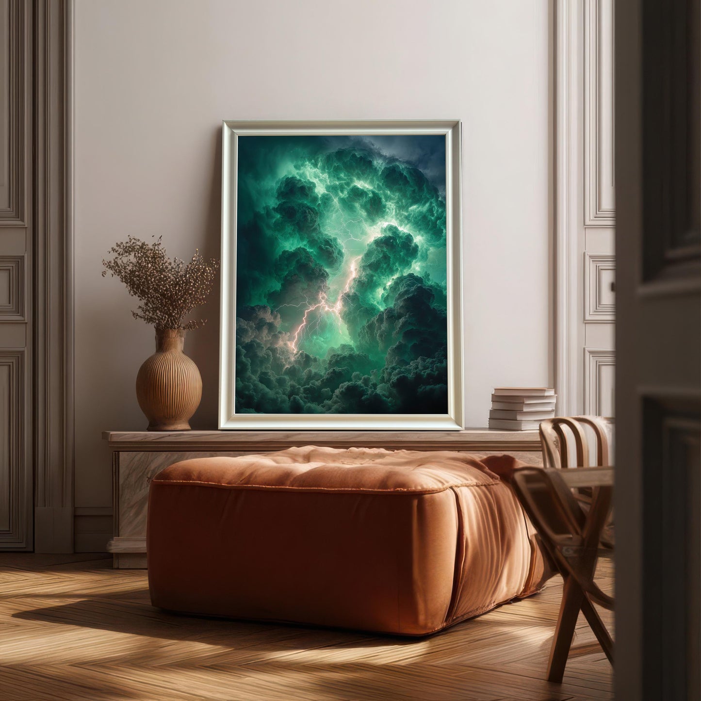 A framed art print of dramatic green and dark storm clouds with lightning resting on a low marble console table behind a large, square, burnt-orange leather ottoman. The frame is White.
