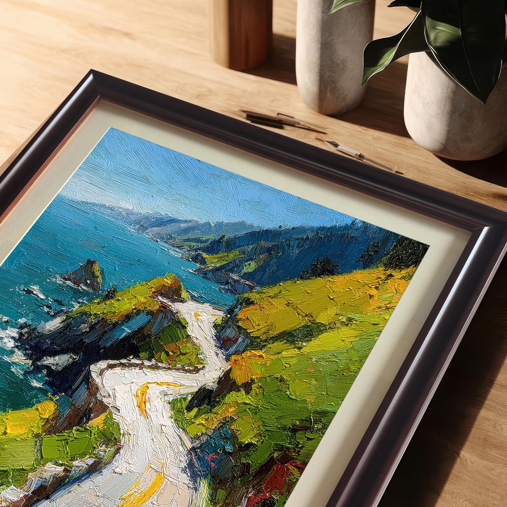 A close-up, angled view of the framed print of the oil painting-style coastal road, resting on a wooden desk. The frame is Coffee (dark brown) with a cream-colored mat. The background shows neutral desk decor and a potted plant.