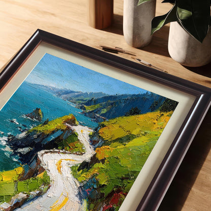 A close-up, angled view of the framed print of the oil painting-style coastal road, resting on a wooden desk. The frame is Coffee (dark brown) with a cream-colored mat. The background shows neutral desk decor and a potted plant.