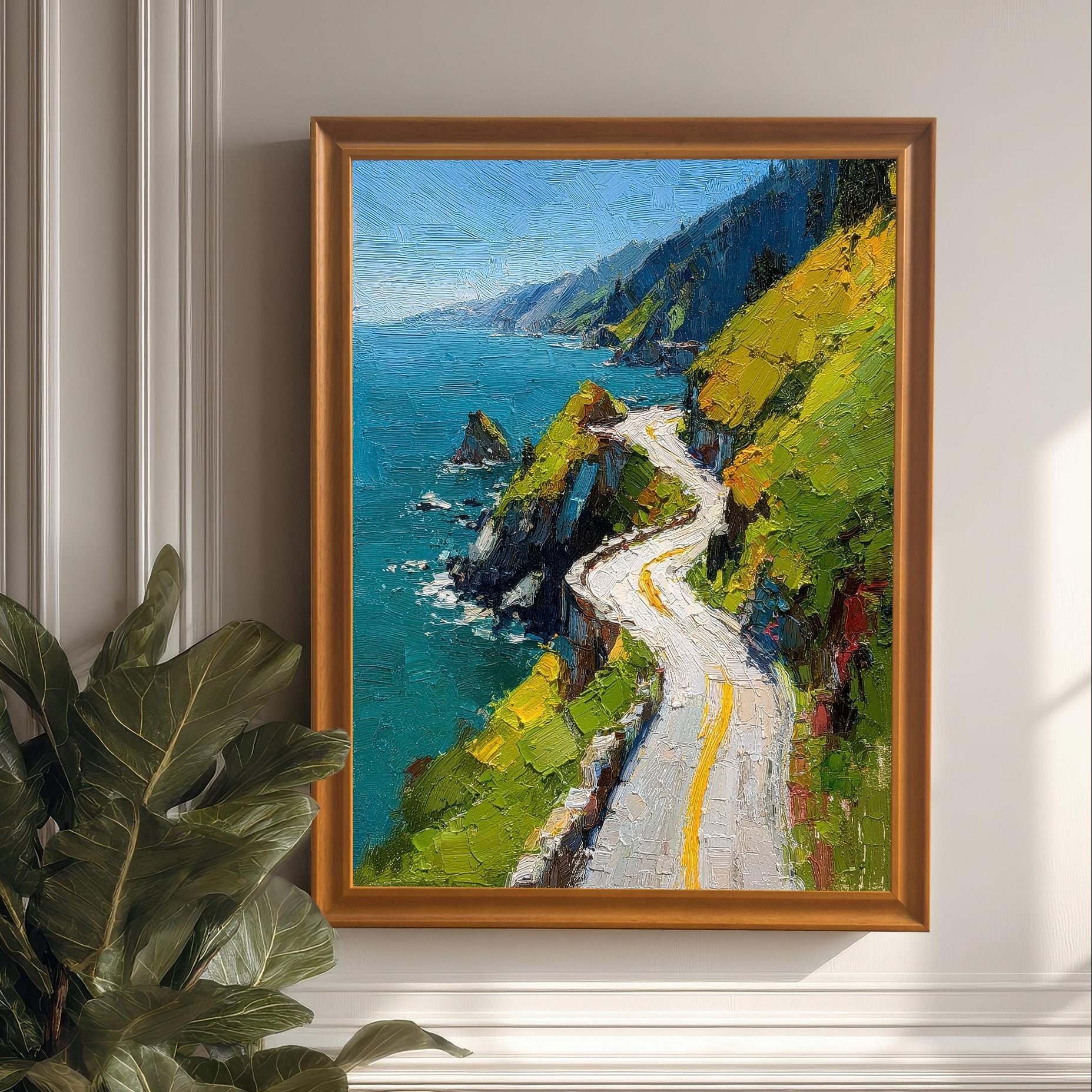 A framed print of a vibrant oil painting of a winding coastal road, hanging on a classic white wainscoted wall, next to a large potted fig-leaf plant. The frame is Orange (light brown wood color).