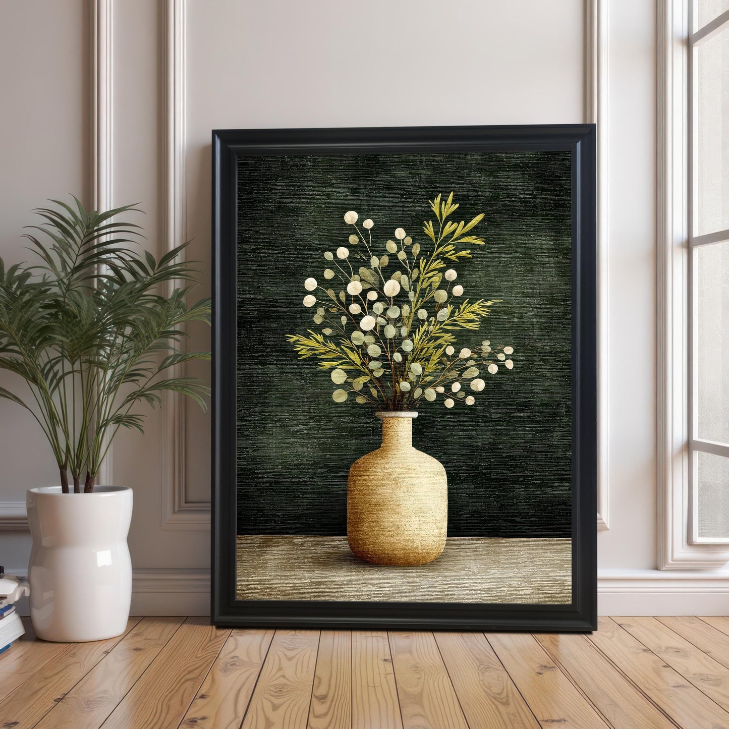 A large framed art print of a vase with foliage, resting on a wooden floor against a wall with white molding. The frame is Black with no mount/border visible. A potted green plant is visible on the left.