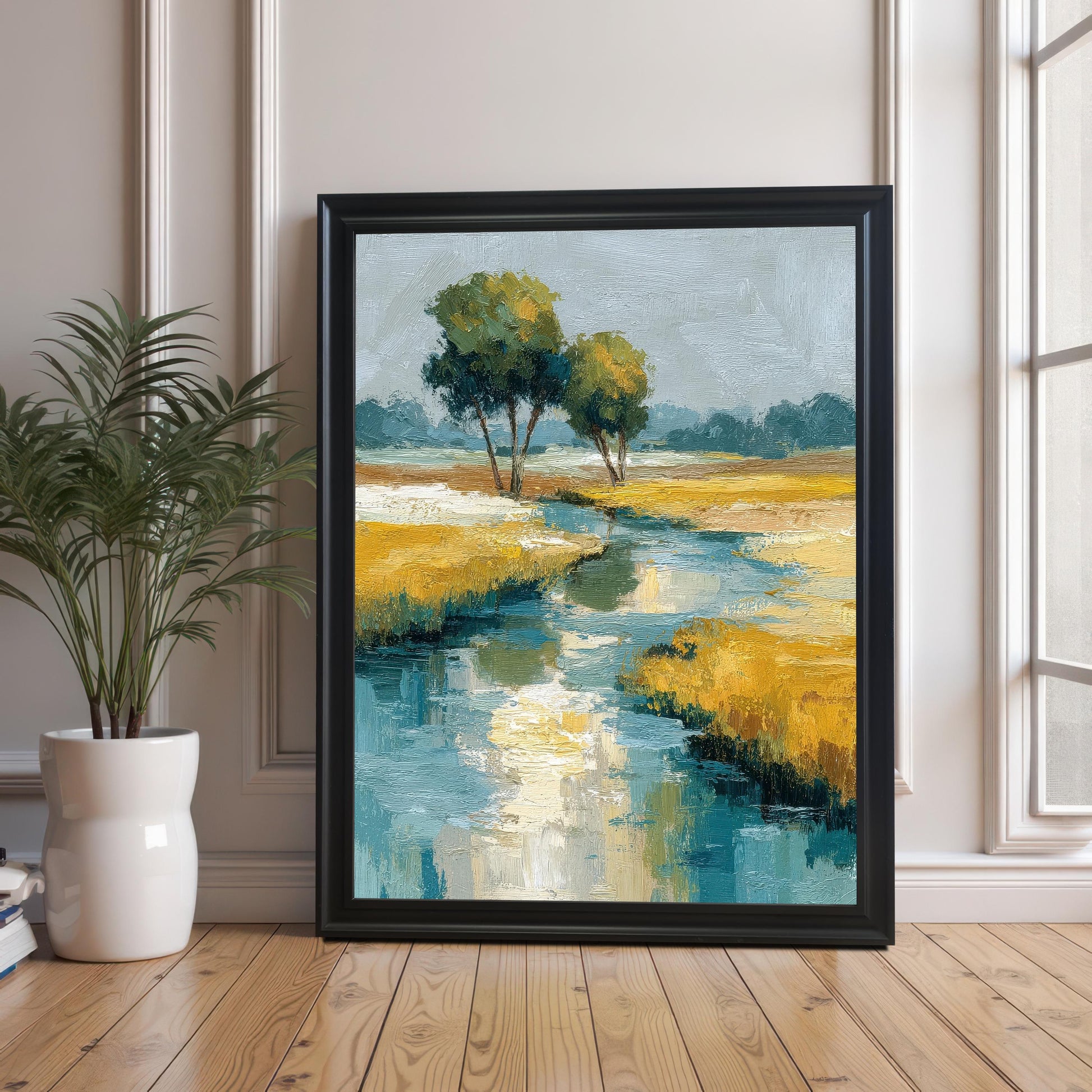 A large framed art print of the abstract landscape painting (featuring a stream, yellow grass, and two trees), resting on a wooden floor against a wall with white molding. The frame is Black with no mount/border visible. A potted green plant is visible on the left.