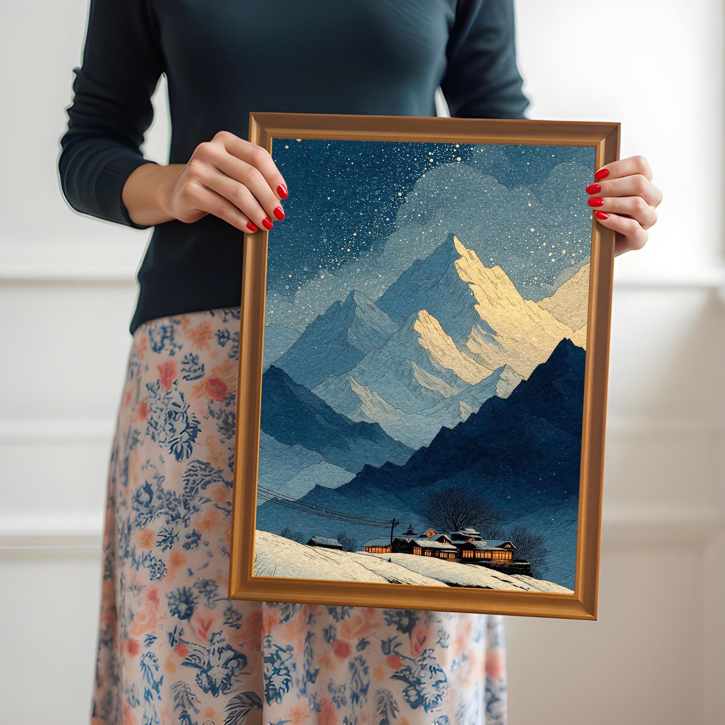 A woman in a black top and floral skirt holds up a framed print of a snowy mountain landscape at night. The art features blue mountains, golden peaks, falling snow/stars, and a cozy cabin with glowing windows. The frame is Orange/Gold wood.