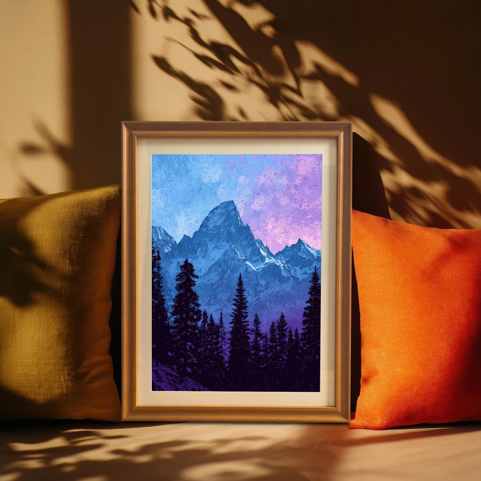 A framed mountain art print with an Orange Frame is propped up between two throw pillows (one olive green, one orange) on a sunny bench. Sunlight casts shadows of leaves or branches onto the wall behind it.