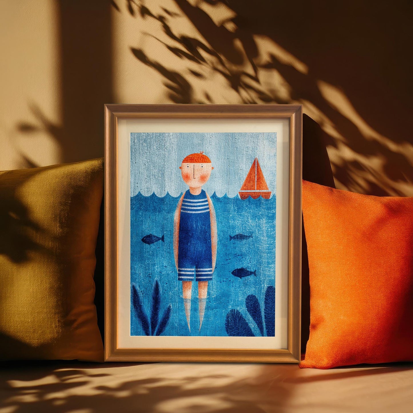 A close-up of the blue and orange nautical art print in a thin gold or light wood frame, leaning against a tan wall between an olive green and an orange throw pillow, with sunlit plant shadows cast across the background.