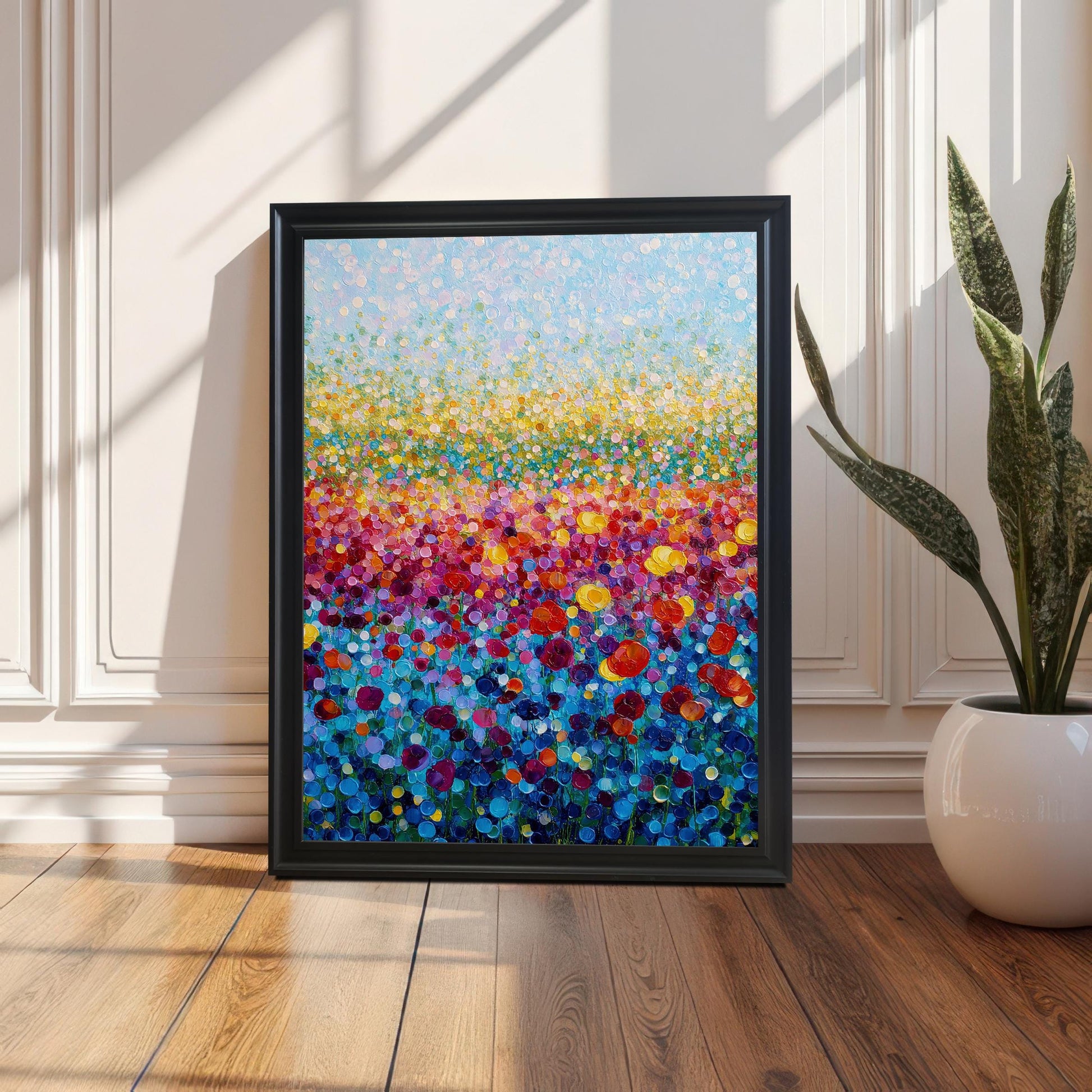 Vibrant abstract flower field print in a simple black frame, leaning against a white paneled wall next to a potted snake plant on a wood floor.