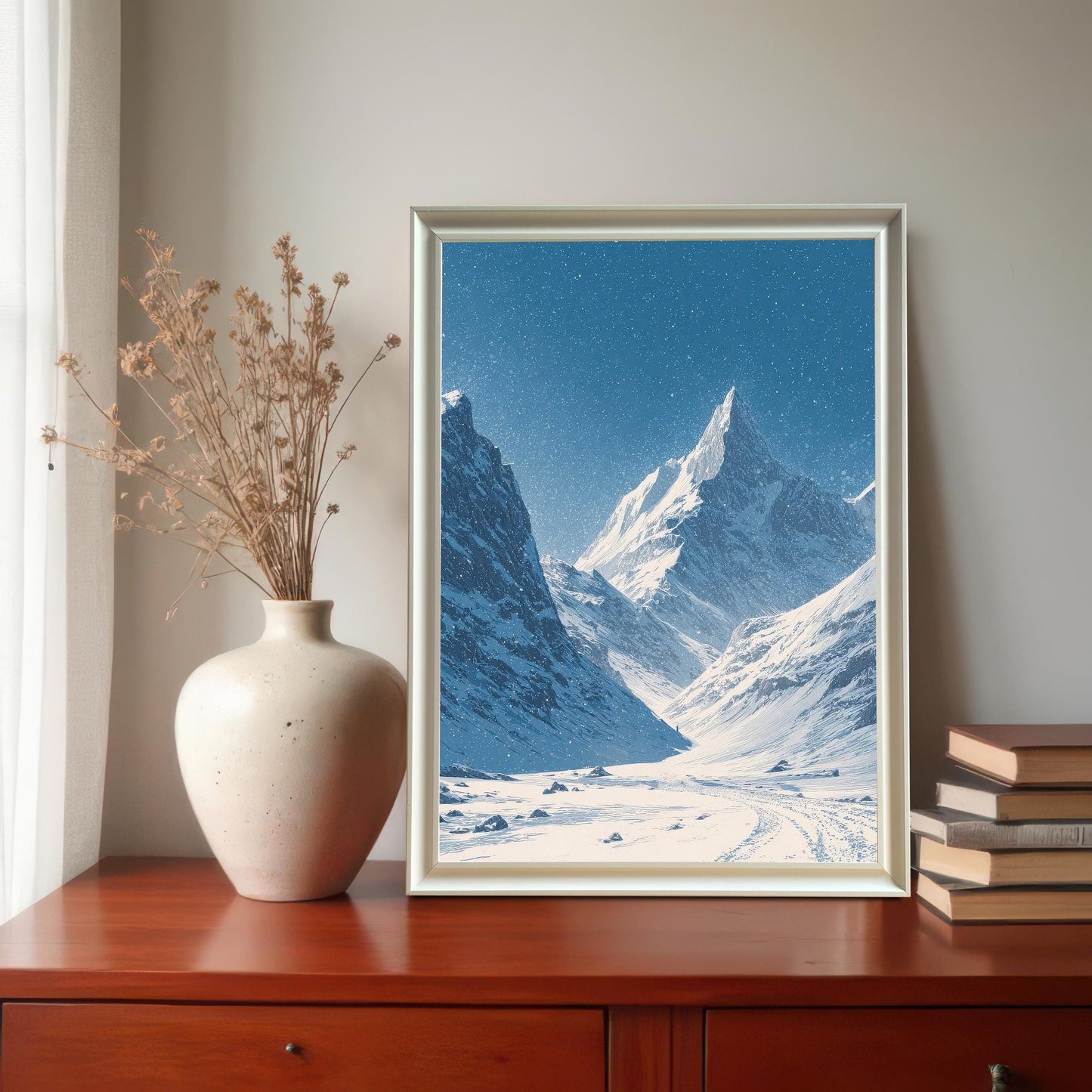 Snowy mountain valley print in a white frame, displayed on a wooden chest of drawers next to a ceramic vase with dried grass and a stack of books.
