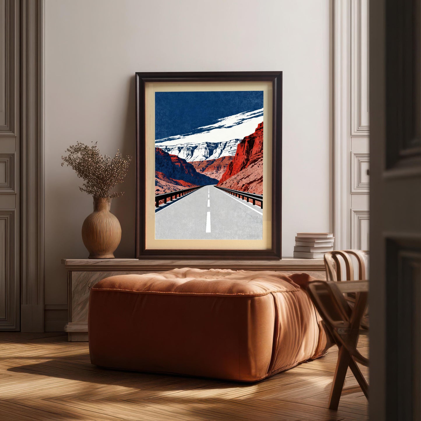 A framed art print with a dark brown frame, leaning against a neutral-colored wall in a styled room. The print is above a large, cushioned, orange-brown ottoman.