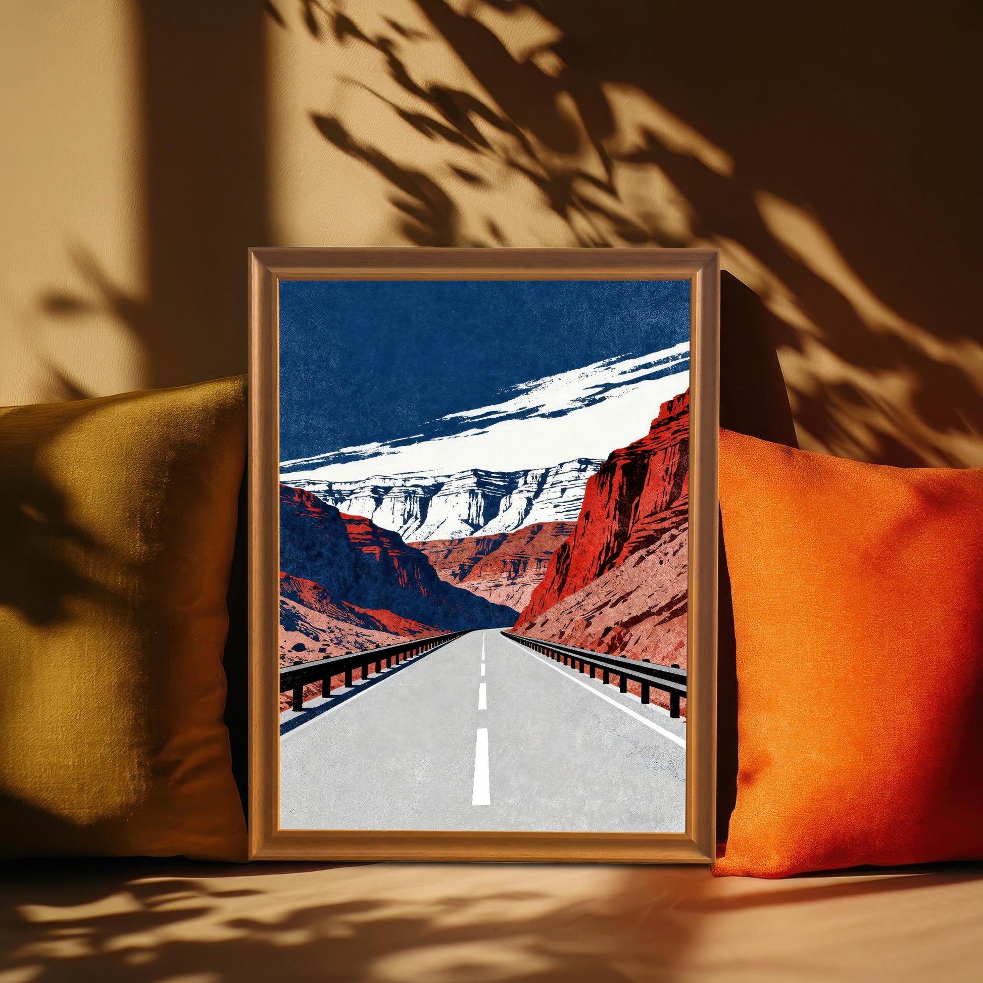 A framed art print with a brown frame, leaning between two pillows on a sofa, with striking shadows cast across the background. The art depicts a long road through canyons.