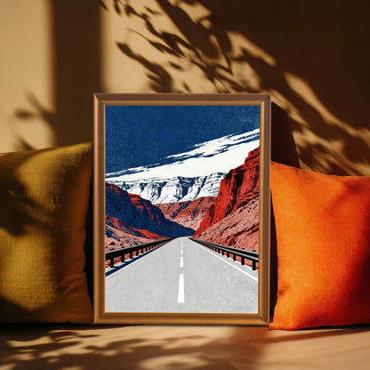 A framed art print with a brown frame, leaning between two pillows on a sofa, with striking shadows cast across the background. The art depicts a long road through canyons.