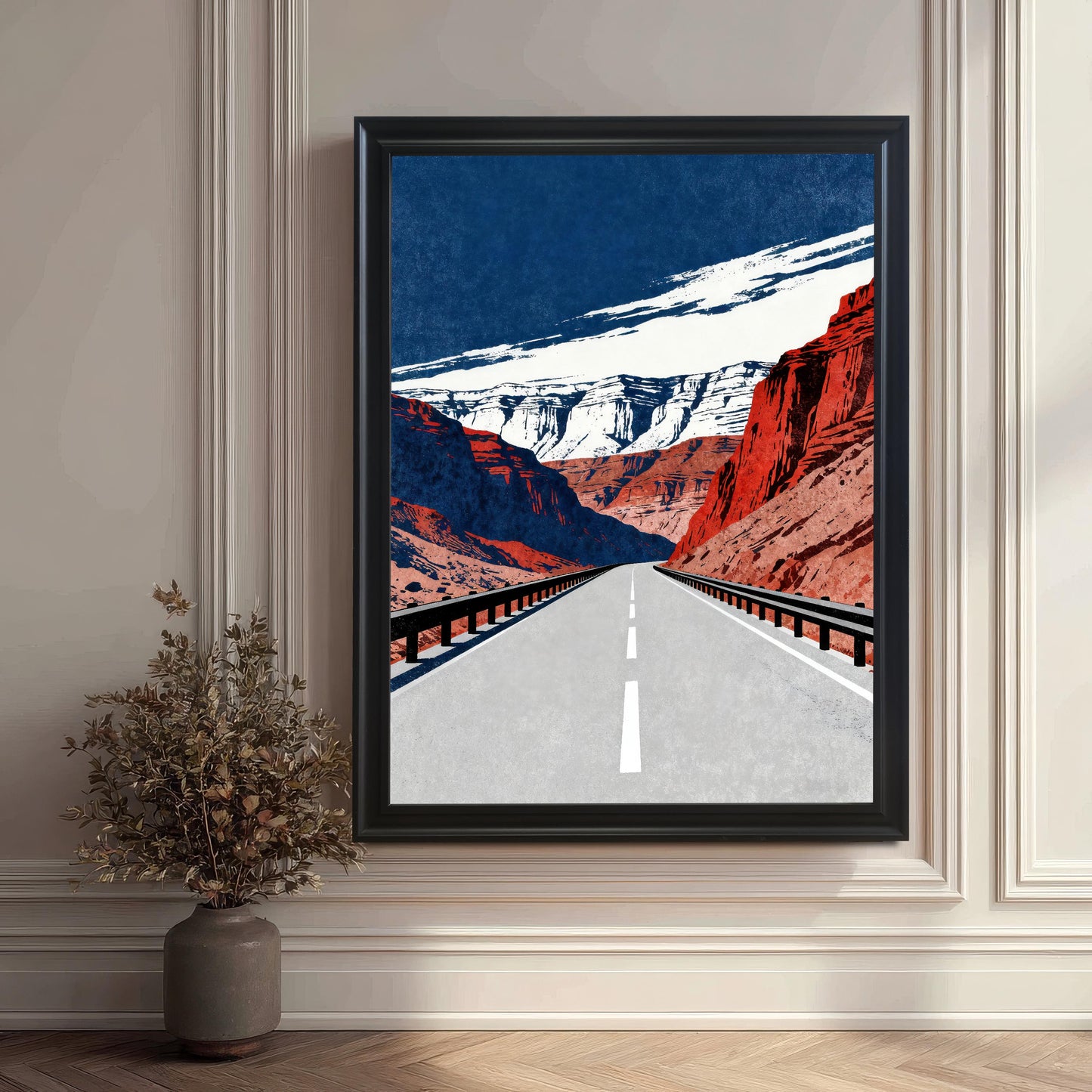 A large, framed art print featuring a road running through red rock canyons, set in a room with elegant white wainscoting and a decorative vase of dried plants on the floor. The frame is black.