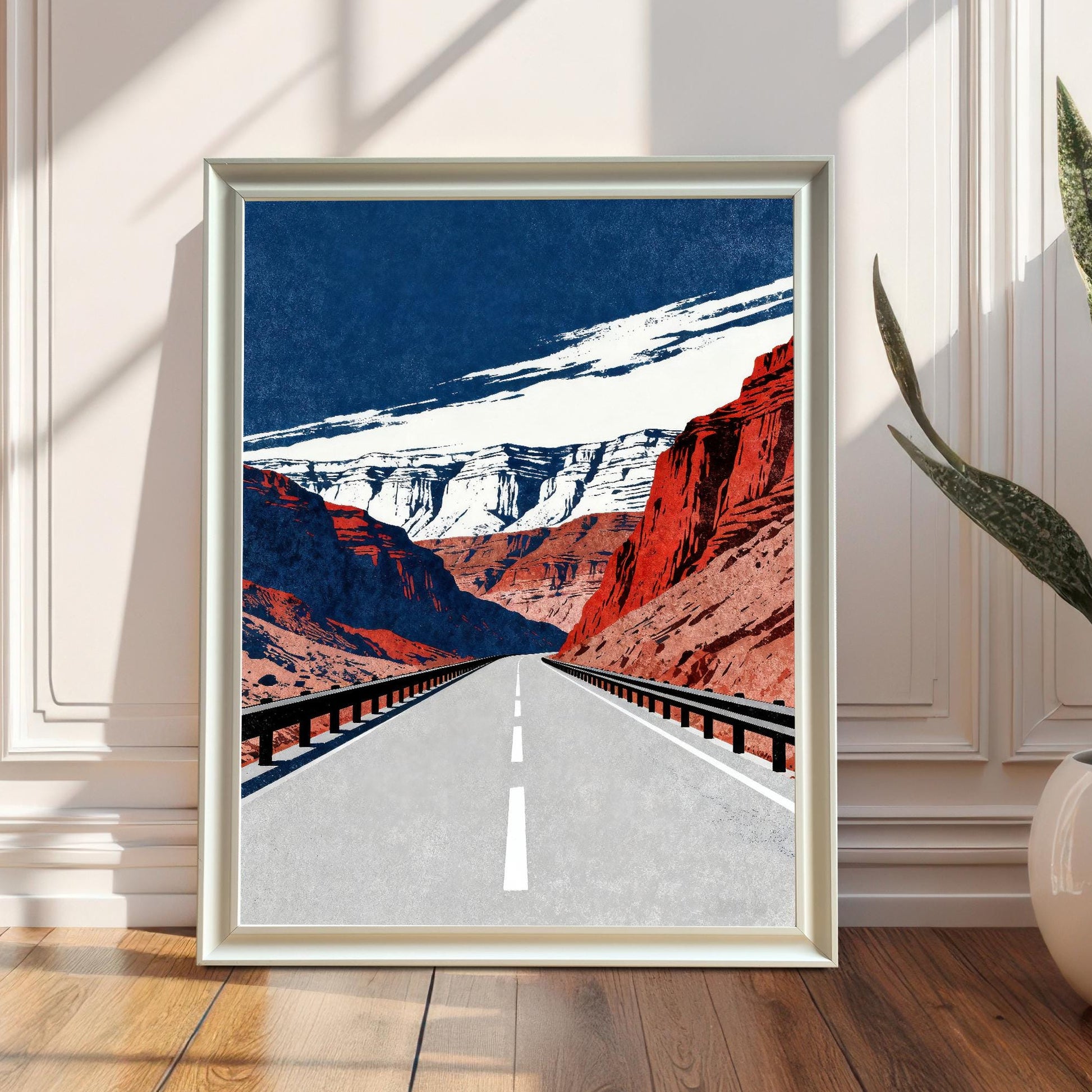 A framed art print with a white frame, leaning against a white wainscoted wall on a wooden floor. The print is bathed in sunlight and shadow, and a large potted snake plant is visible on the right.