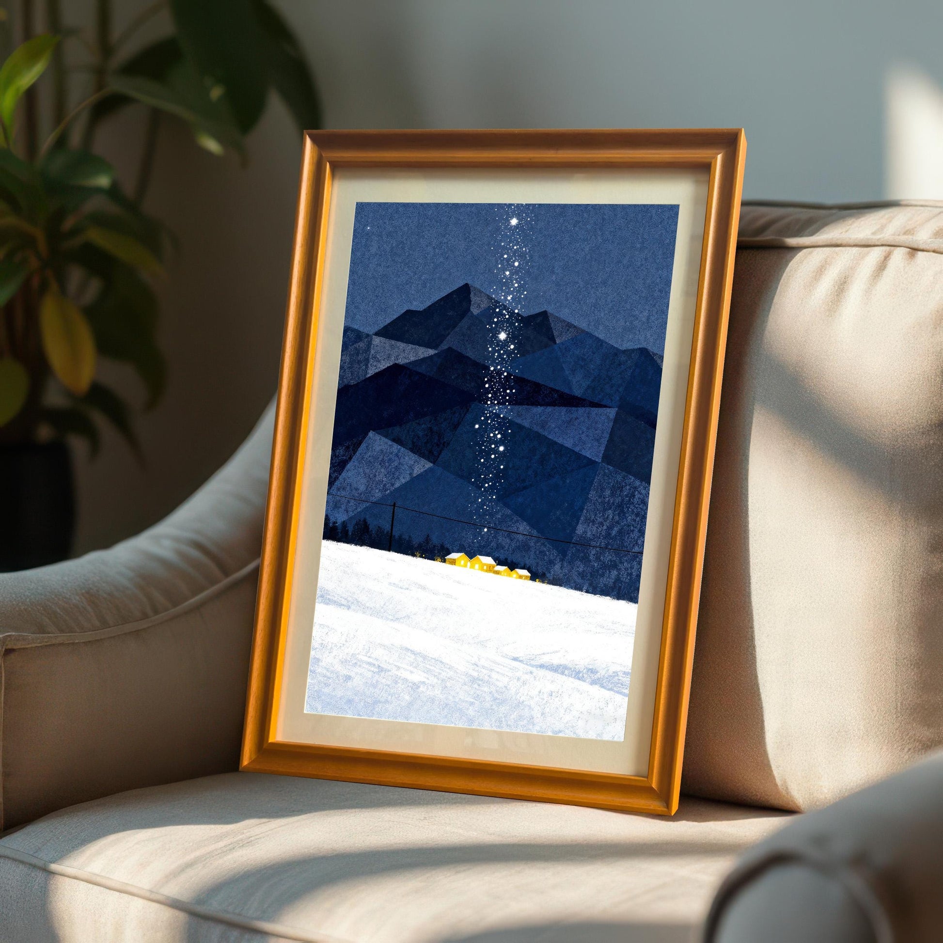Framed art print in an Orange Frame with a mount, featuring dark blue geometric mountains, snow, and a starry trail, resting on a beige armchair indoors with a plant visible in the background.