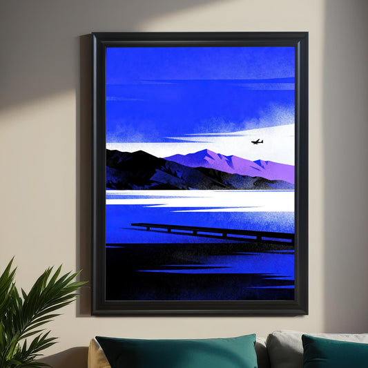 Framed modern art print with a black frame hanging above a bed. The artwork is a stylized landscape featuring vibrant blue sky and water, black and purple mountains, and a small airplane in the distance. The room has dark green and gold curtains, a houseplant, and teal accent pillows.