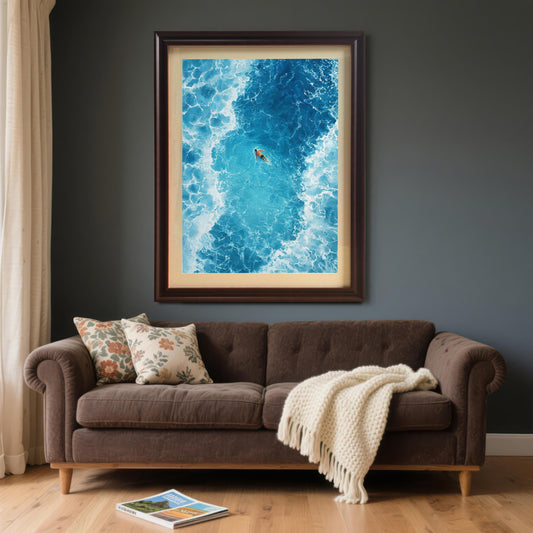 The framed ocean art print in a Coffee/dark brown frame with a mat (mount), hanging on a dark grey wall above a brown velvet sofa with throw pillows and a white blanket.