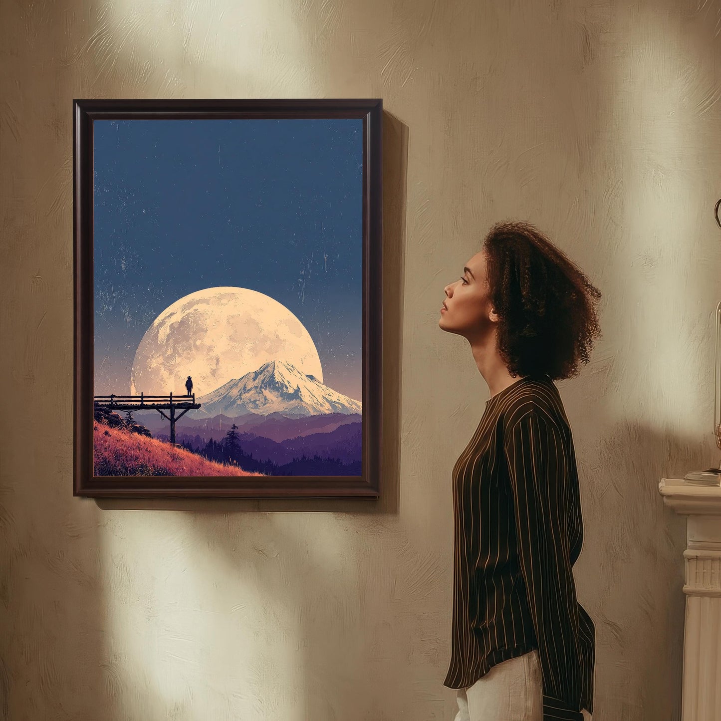 Landscape art print in a dark wood frame, featuring a large moon over a mountain and a figure on a bridge, hanging on a textured wall with a woman looking at it.