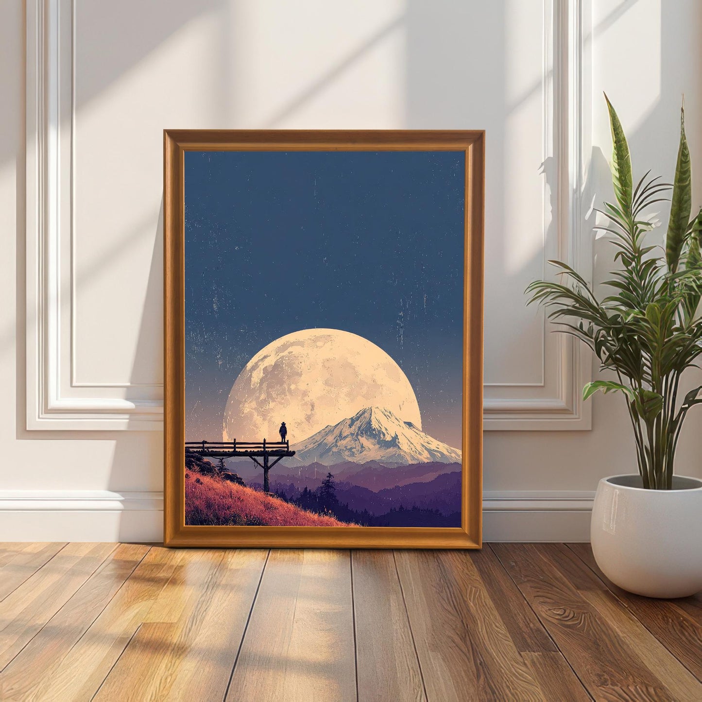 Landscape art print in an orange/light brown wood frame, leaning on a white decorative wall next to a tall potted plant.