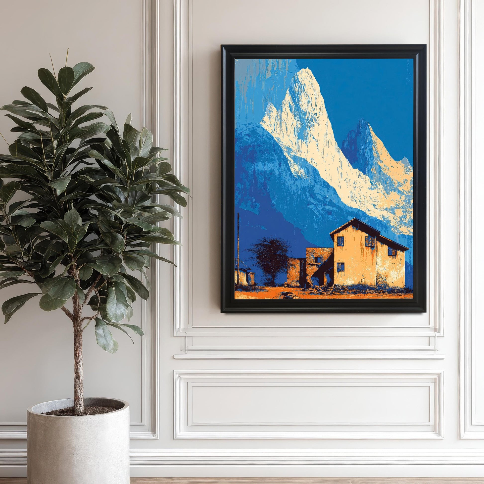 Large landscape art print in a black frame, featuring stylized blue and white mountains and an orange house, hanging on a white decorative wall next to a tall potted plant.