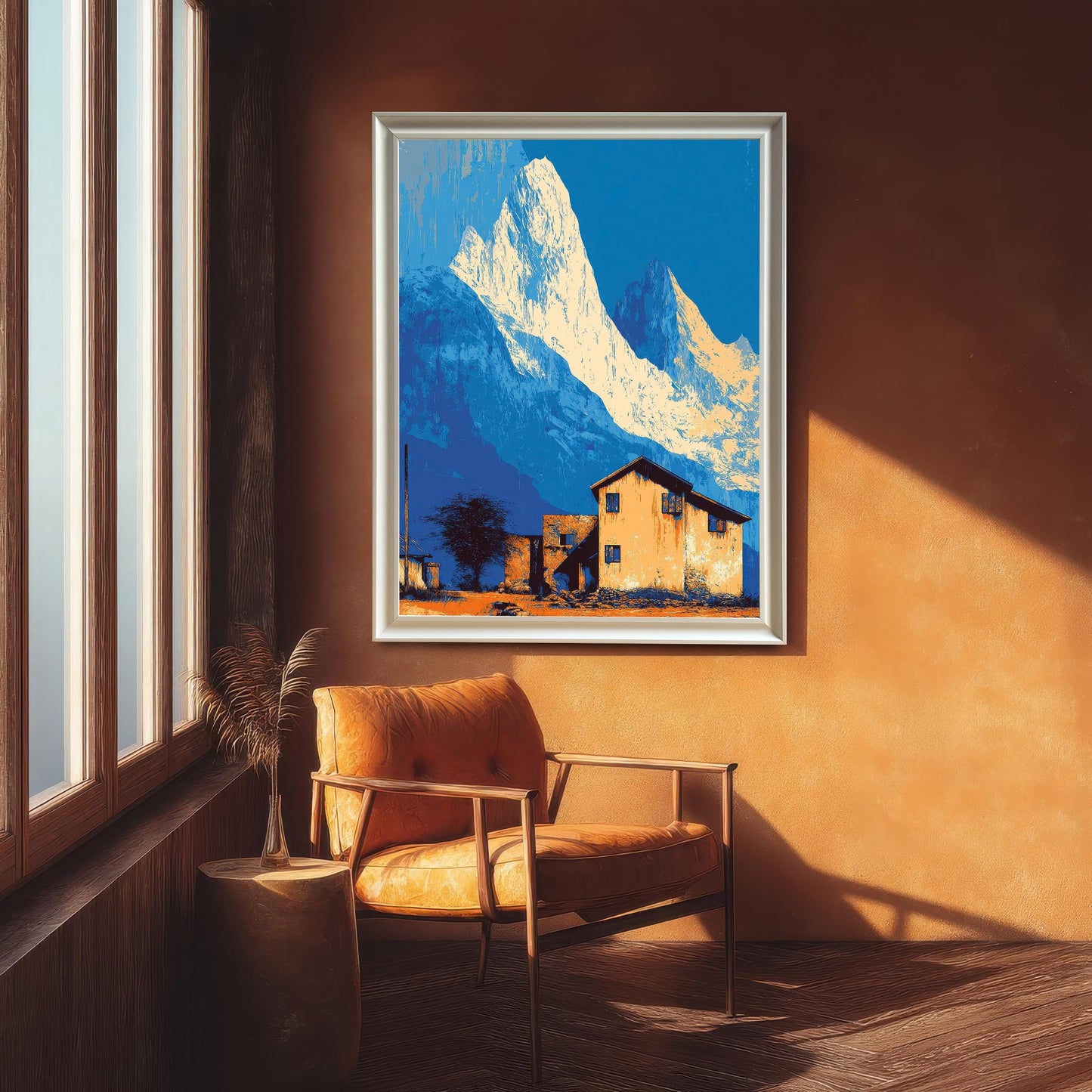 Large landscape art print in a white/silver frame, featuring stylized blue mountains and an orange house, hanging in a room with an orange wall above a leather armchair.