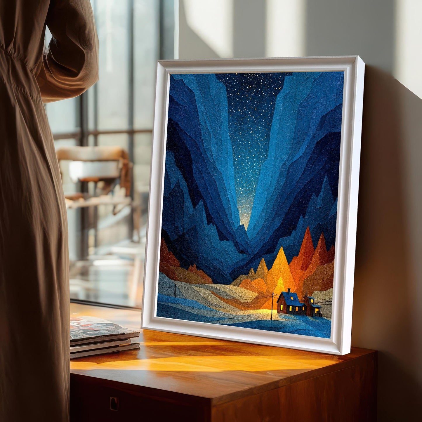 The geometric mountain art print in a White Frame / No Mount (full bleed print) leaning on a wooden chest near a window, with a person in a neutral-colored dress standing in the foreground.