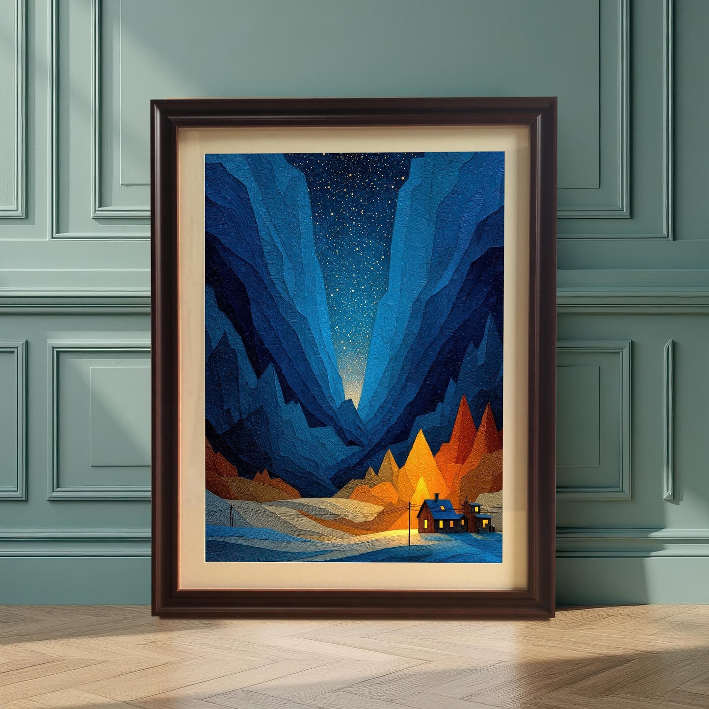 The framed geometric art print, featuring a snowy mountain valley and an illuminated cabin, leaning on a wooden floor against a paneled wall painted dark teal. The frame is a dark, Coffee color with a mat/mount.
