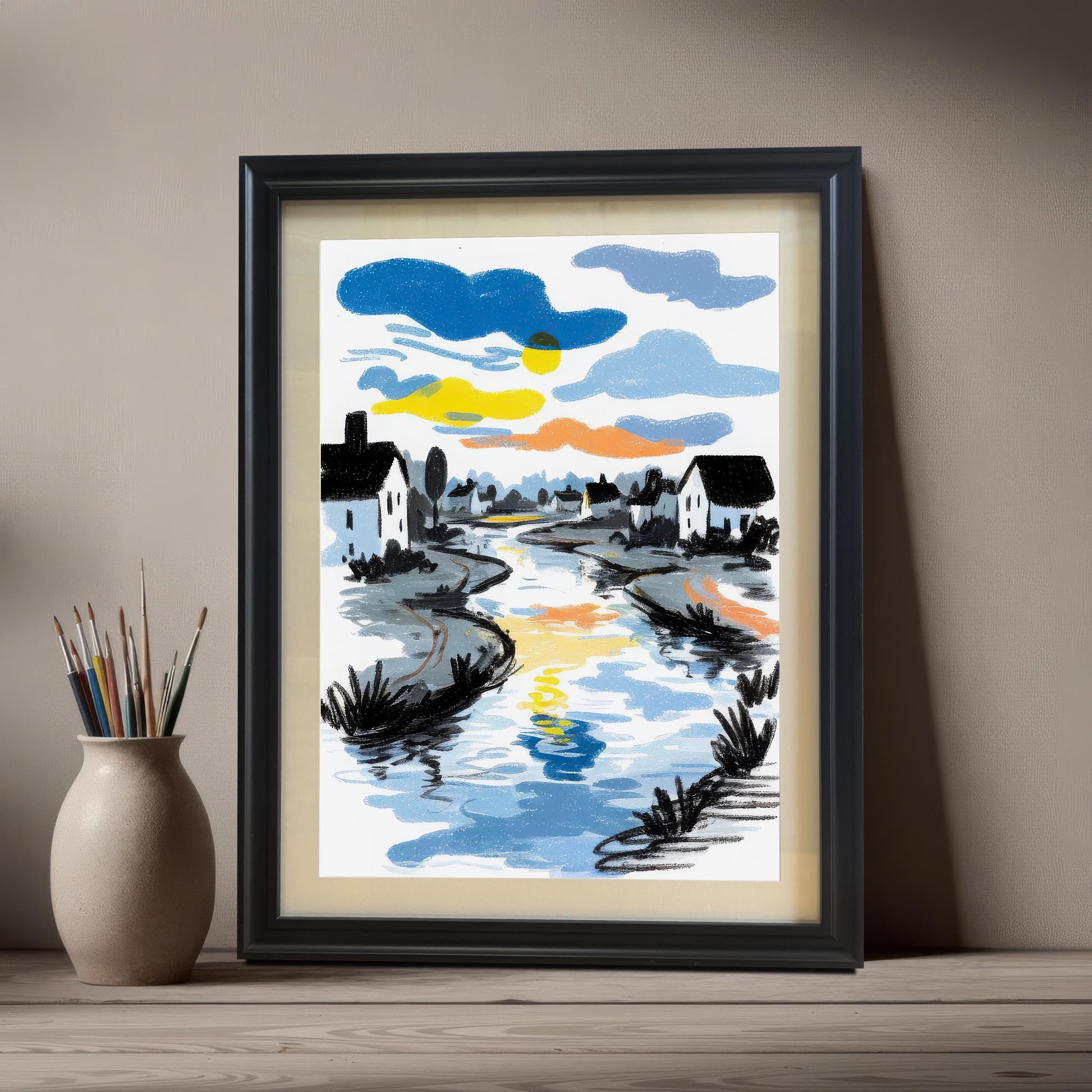 The river art print in a Black Frame & Mount (with a light mat) leaning on a light wooden table next to a vase holding paintbrushes.