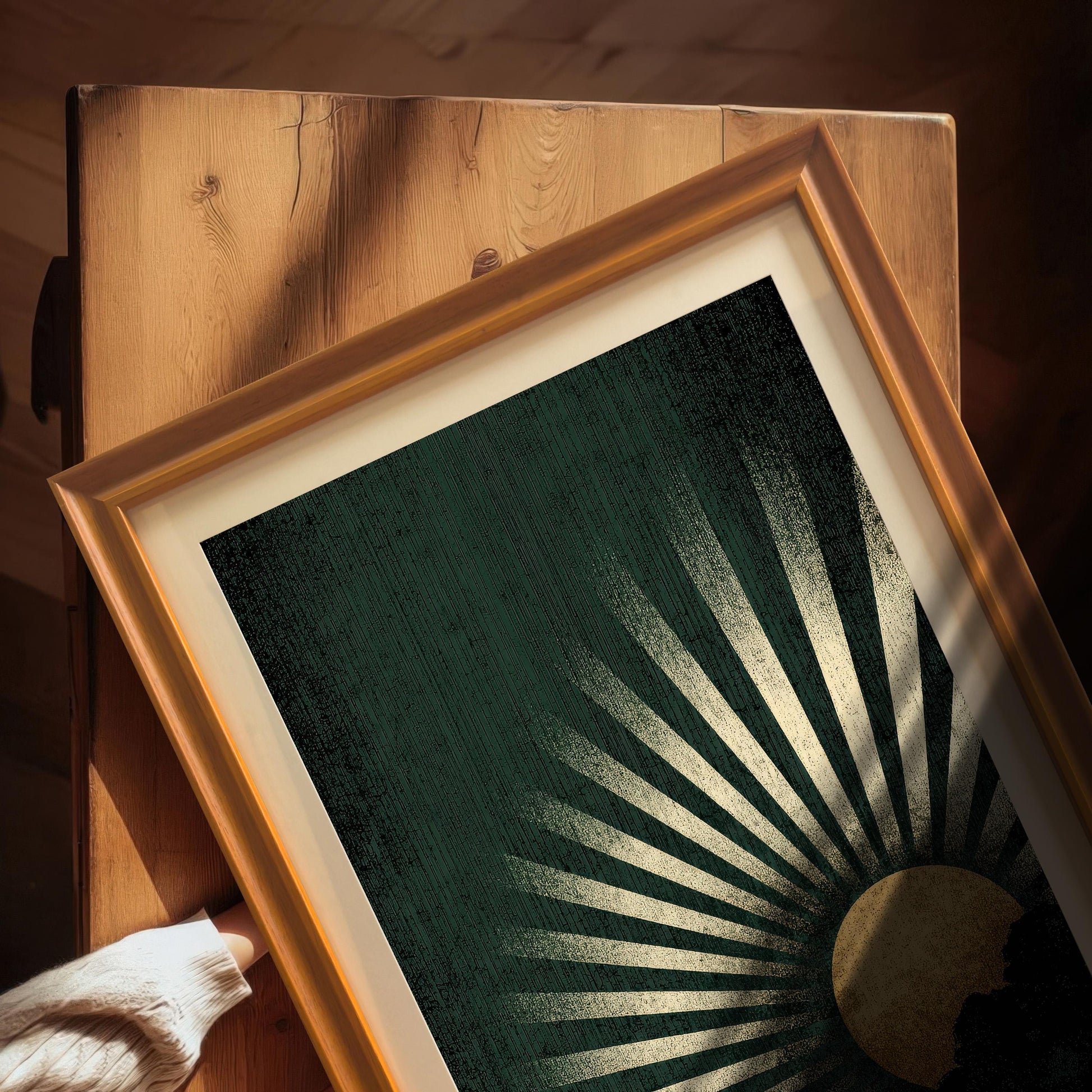 Overhead close-up view of the dark green and gold sunburst art print in an Orange frame with a mount, resting on a wooden surface. A hand in a cream sweater is visible in the corner.