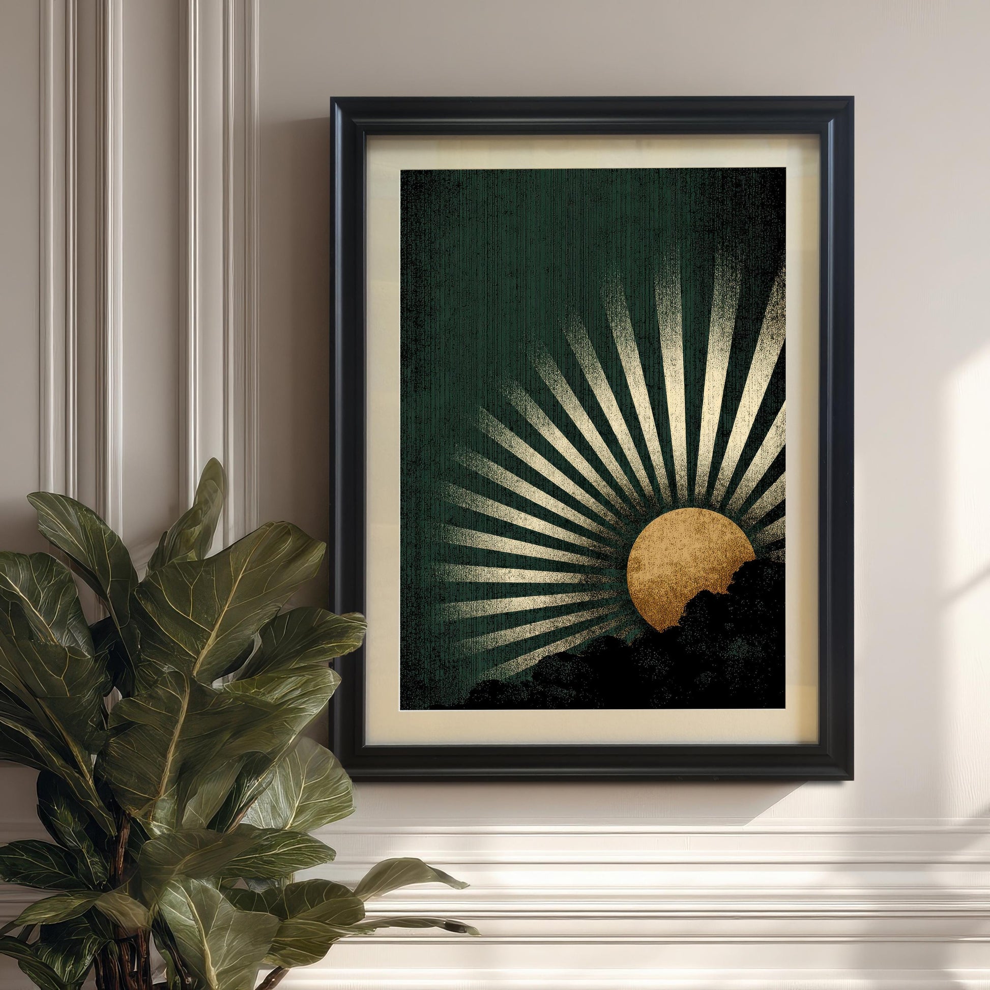 The dark green and gold sunburst art print in a Black Frame with a mount, hanging on a paneled white/cream wall. A large fiddle leaf fig plant is visible in the lower left corner.