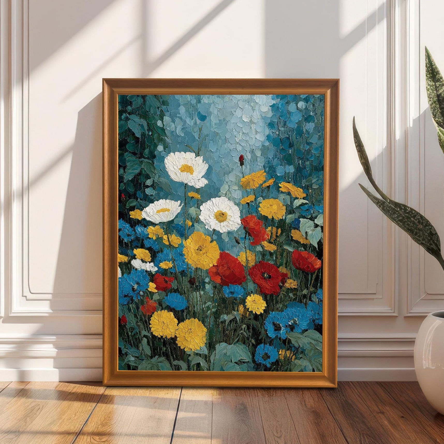An orange-wood framed floral art print leaning against a white paneled wall on a polished hardwood floor, next to a white potted plant in a sunlit room.