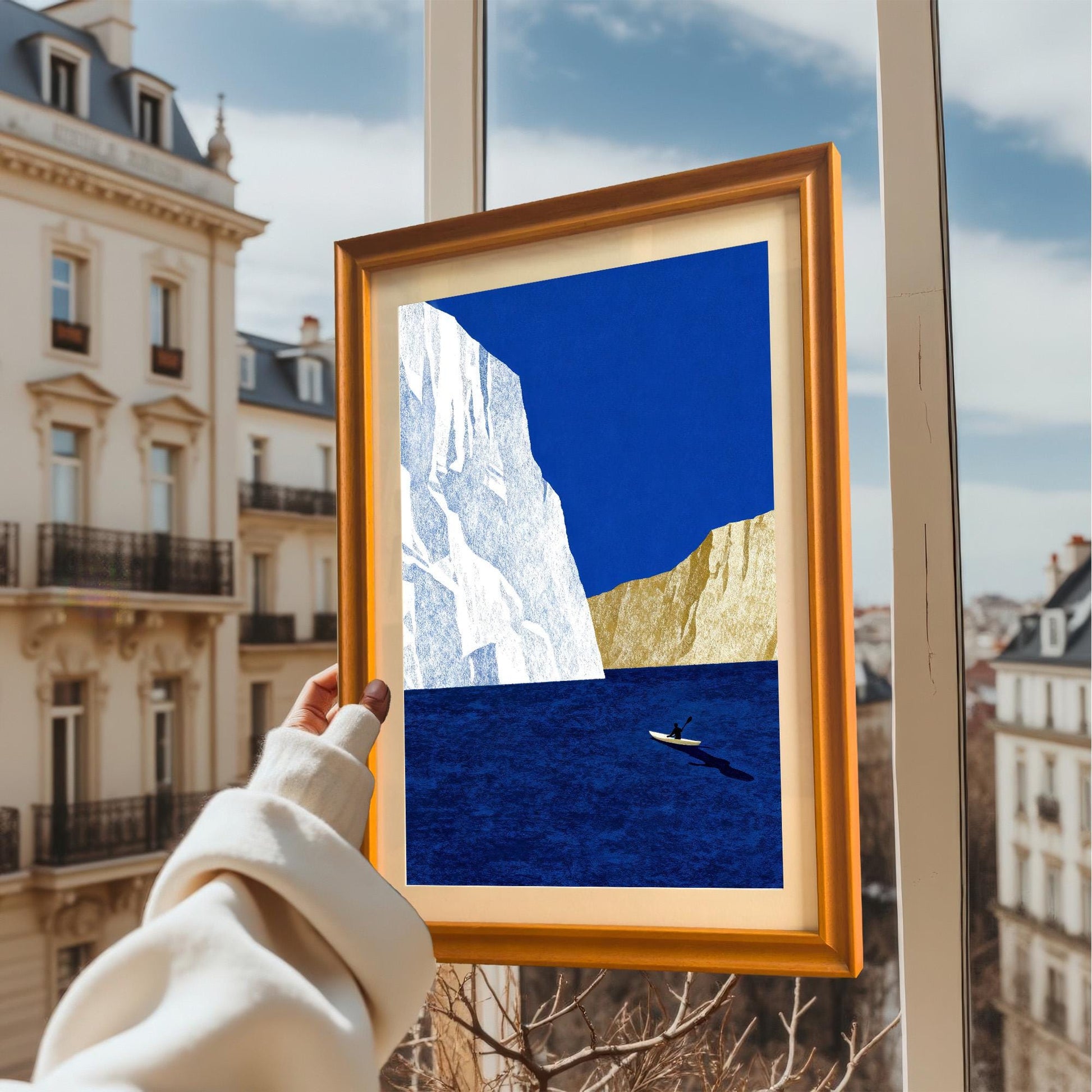 A hand holding an orange wood-framed print of a lone kayaker by an iceberg against a blurred city background through a window.