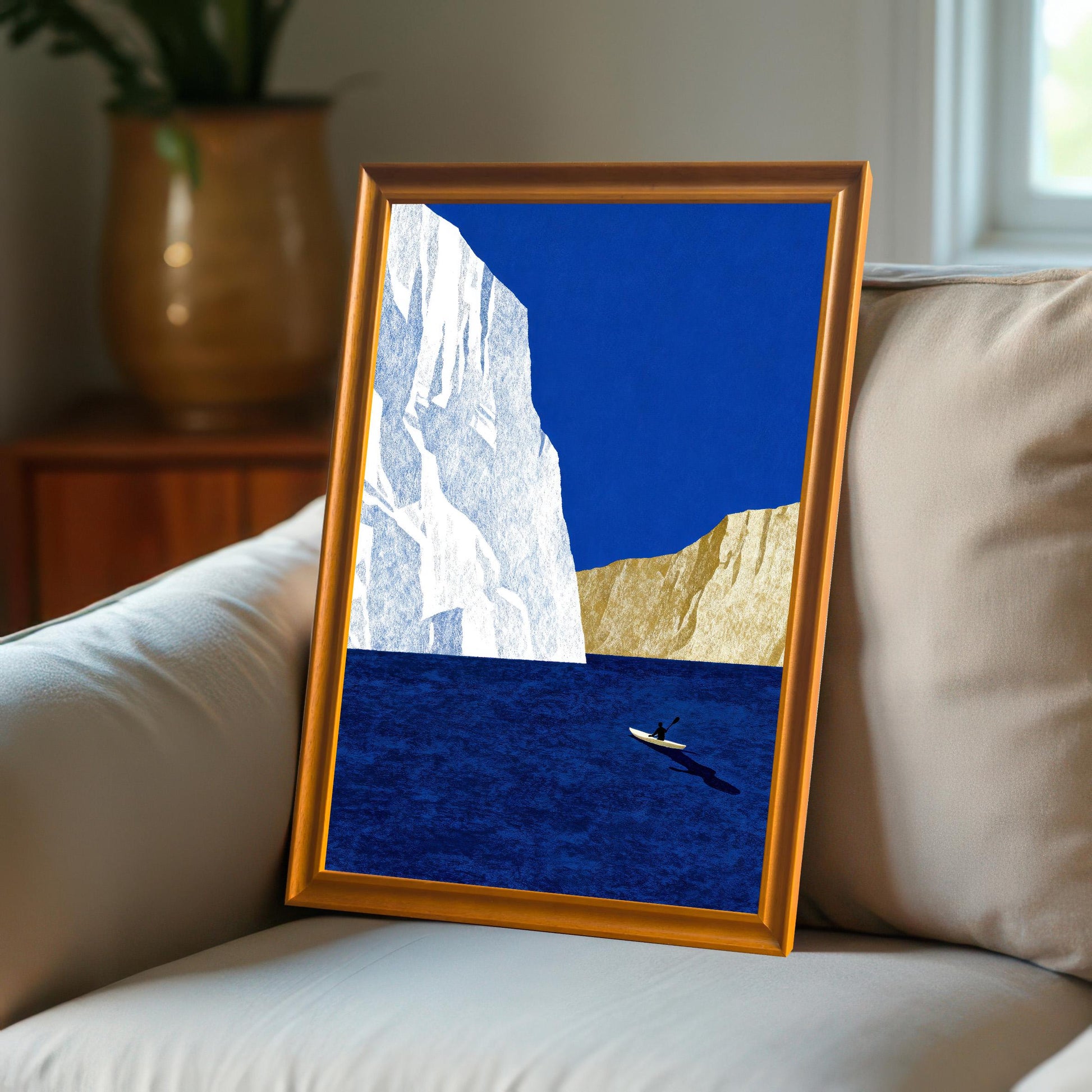 An orange wood-framed minimalist sea print leaning against a beige cushion on a couch in a cozy, sunlit interior.
